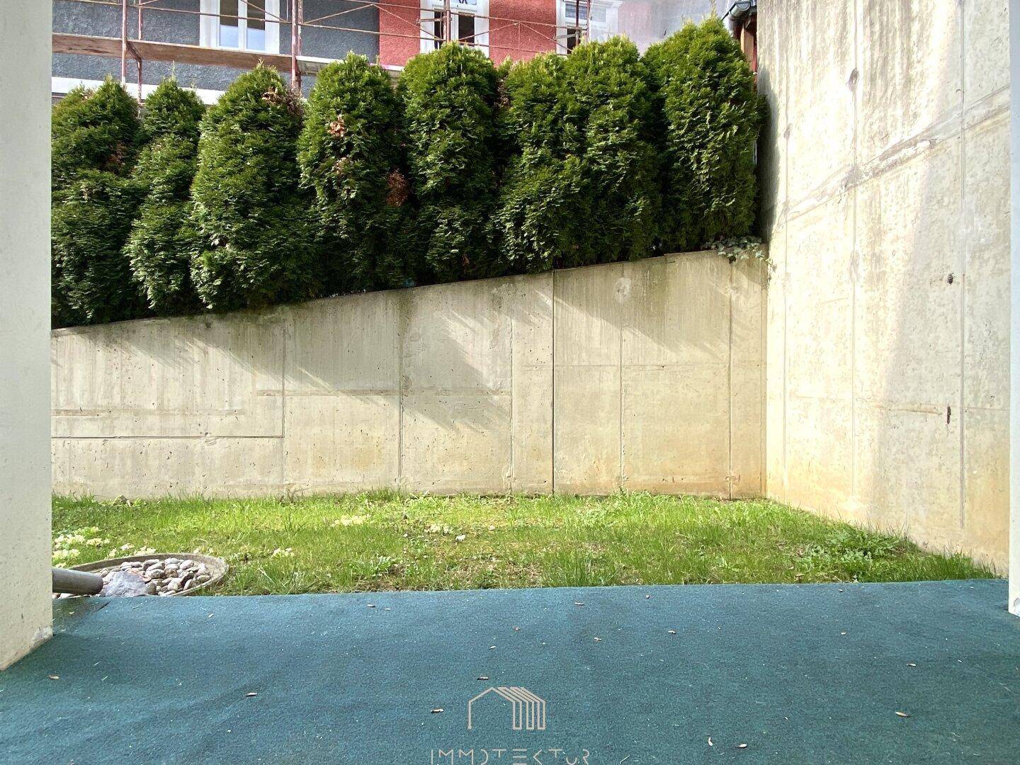 Kleiner Gartenbereich mit Rasenfläche und Hecke, umgeben von einer Betonmauer.