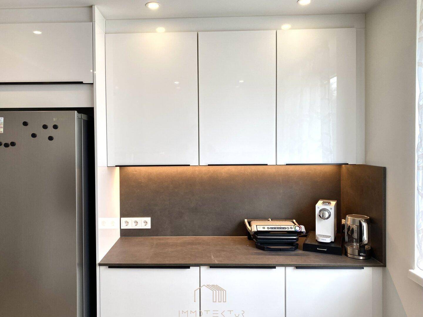 Moderne Küche mit weißen Hochglanzschränken, integriertem Kühlschrank und dunkler Arbeitsplatte.