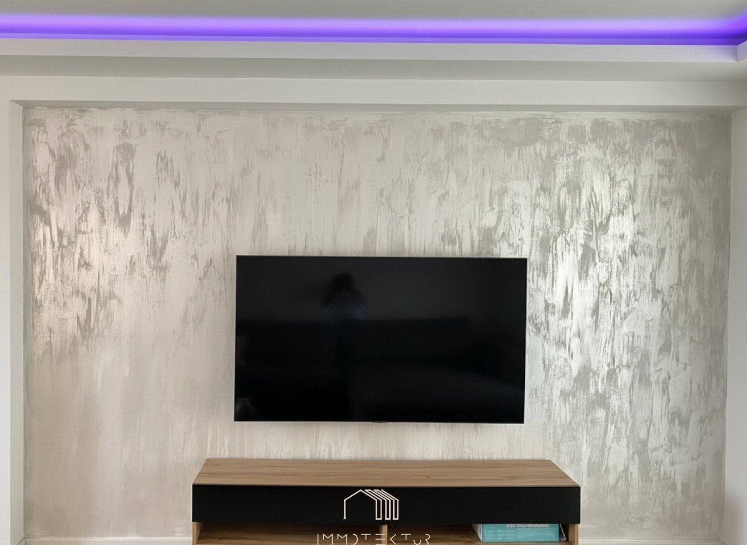 Wohnzimmerwand mit modernem Strukturputz und wandmontiertem Fernseher, beleuchtet durch indirektes Licht.