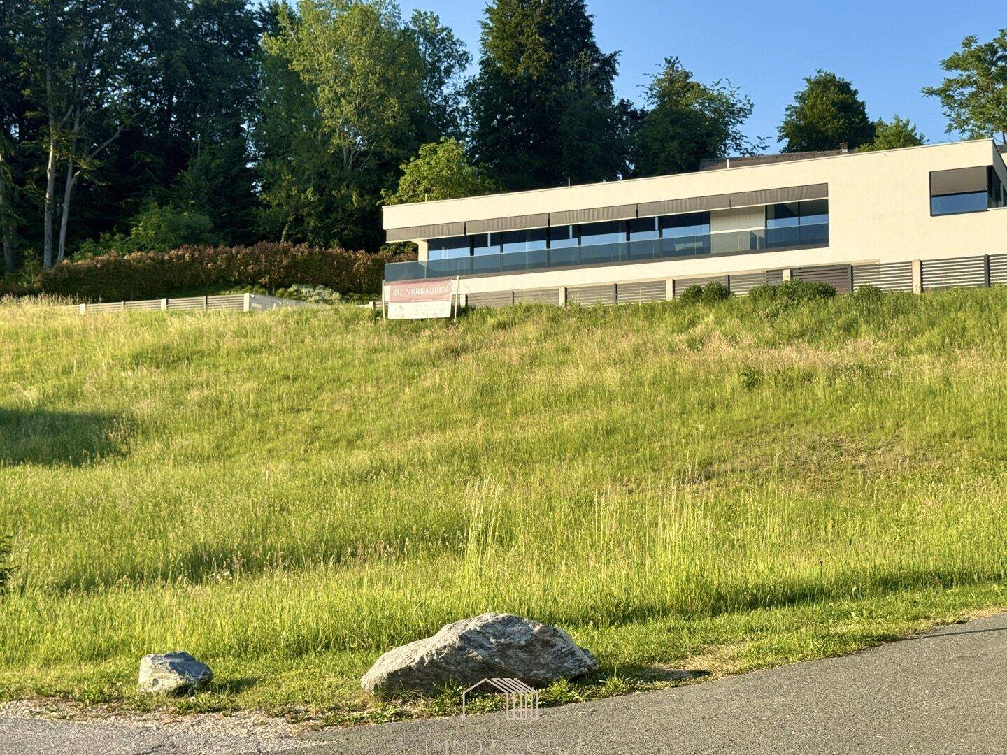 Moderne Architektur auf einem grünen Hügel mit einem "Zu verkaufen"-Schild im Vordergrund.