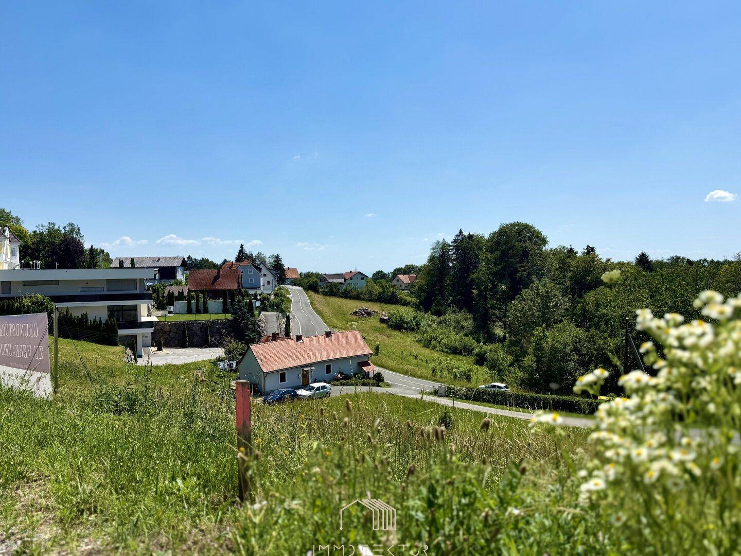 Weitläufiger Blick über eine ländliche Wohngegend mit Häusern, Straße und viel Grün unter blauem Himmel.