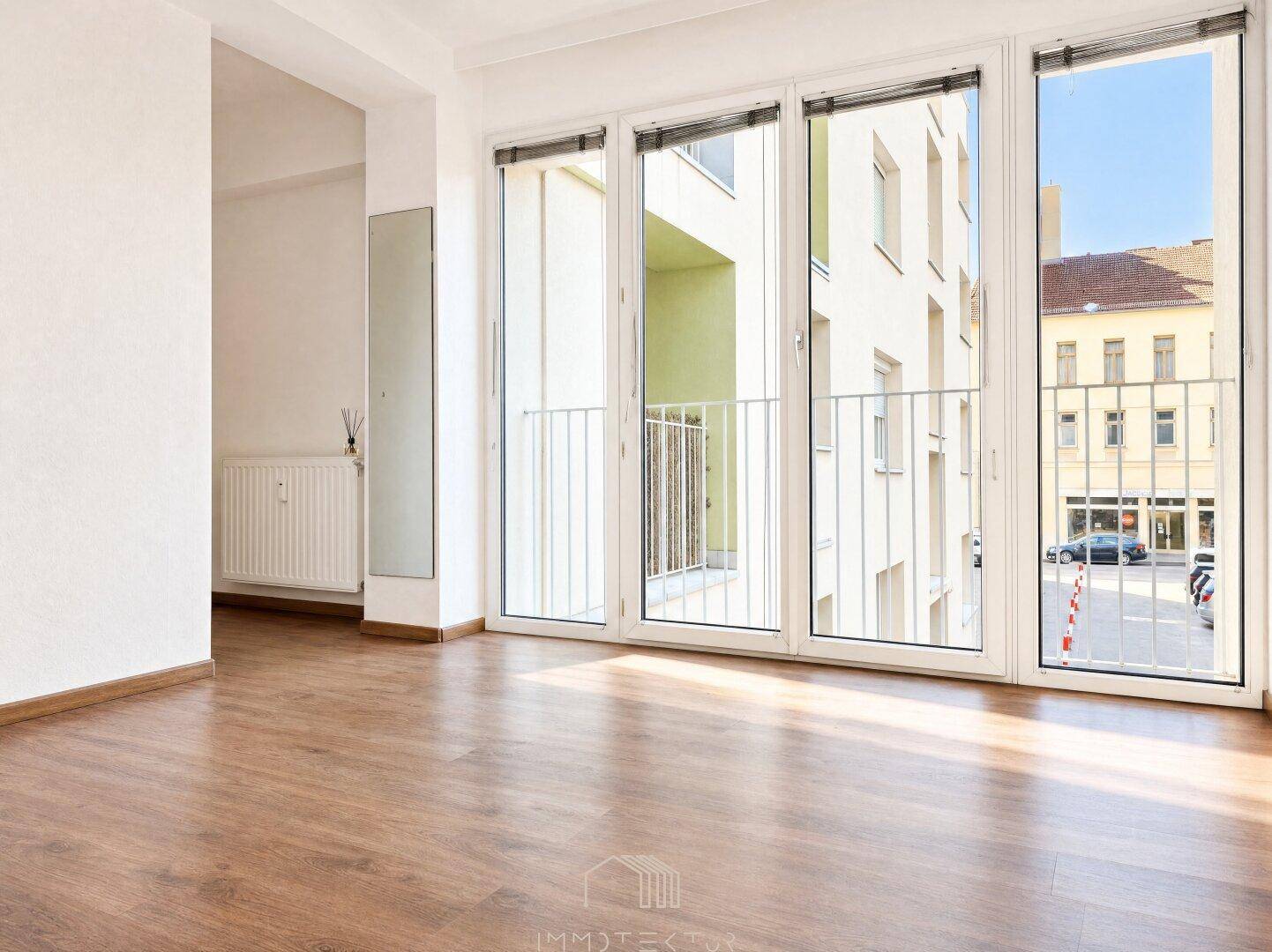 Geräumiges Wohnzimmer mit Laminatboden und großen Fenstern, die viel Tageslicht hereinlassen.