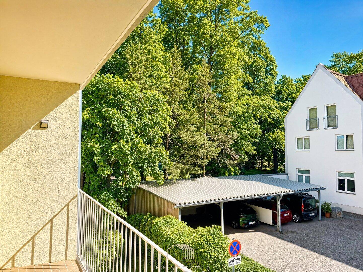 Balkon mit Blick ins Grüne und auf einen überdachten Parkplatz, umgeben von Bäumen.