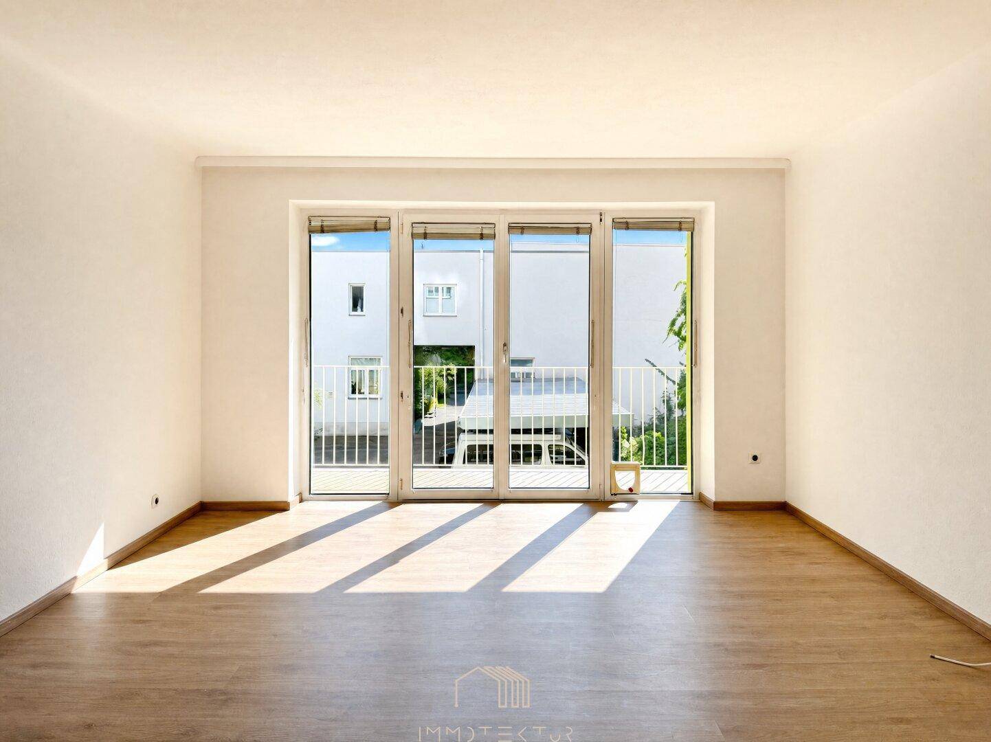 Leeres Wohnzimmer mit Laminatboden und großen Fenstern, die viel Sonnenlicht hereinlassen.