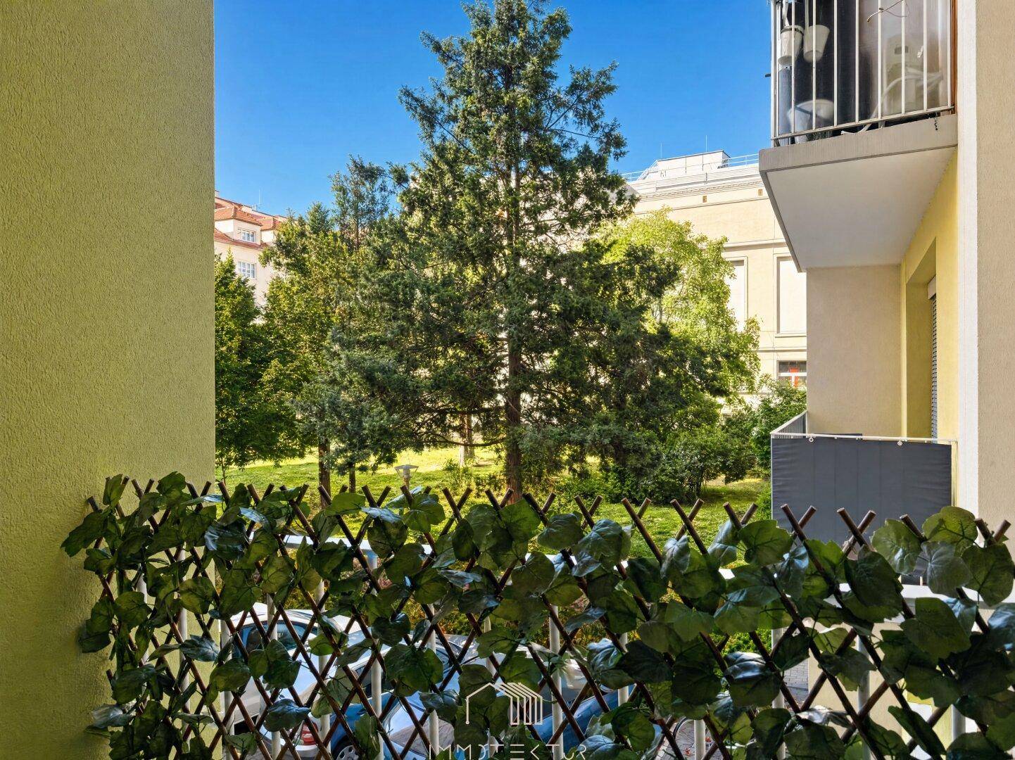 Balkon mit grüner Sichtschutzwand und Blick auf Bäume sowie umliegende Gebäude.