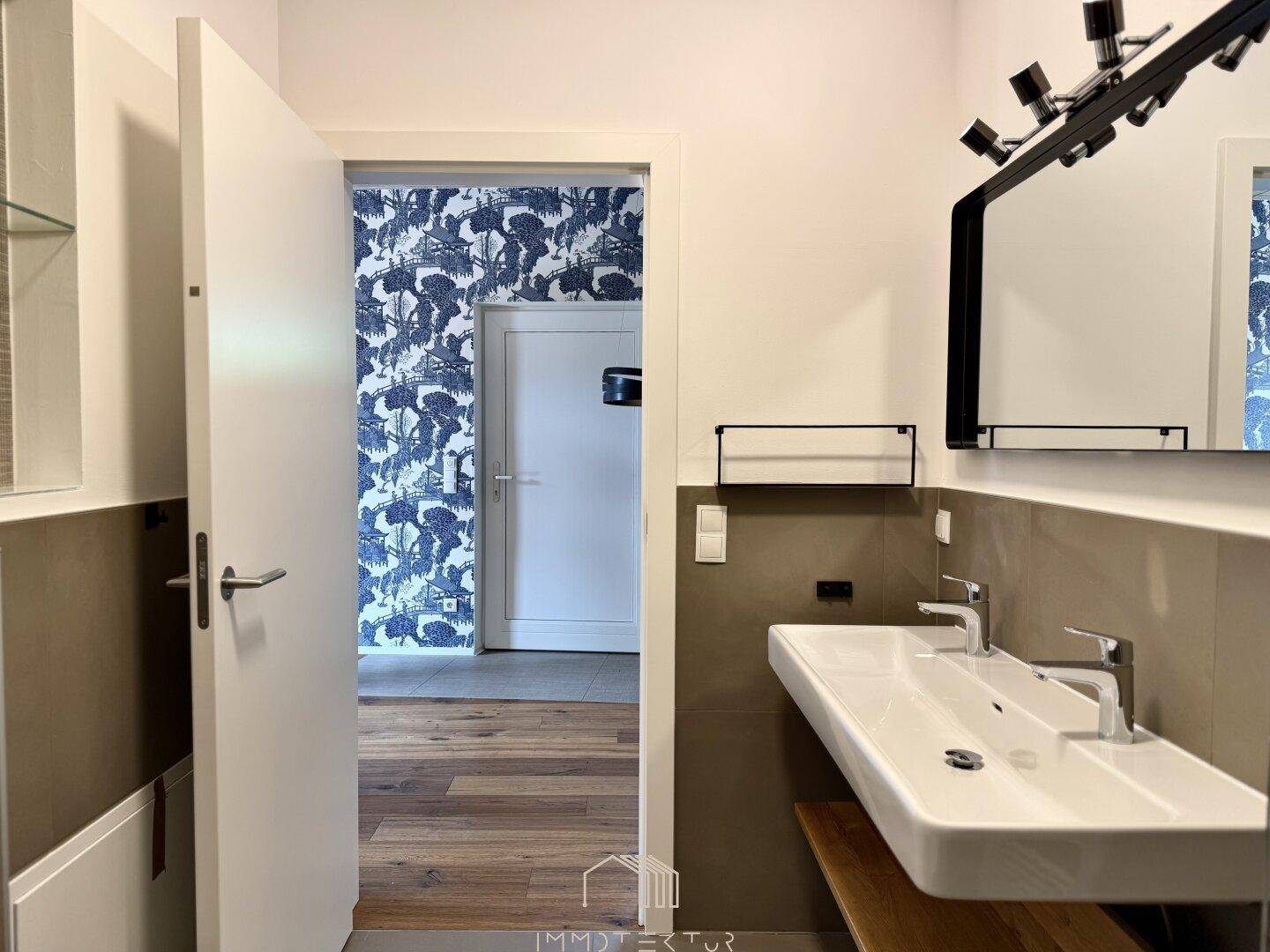 Badezimmer mit Doppelwaschbecken und Blick auf den angrenzenden Raum mit gemusterter Tapete.