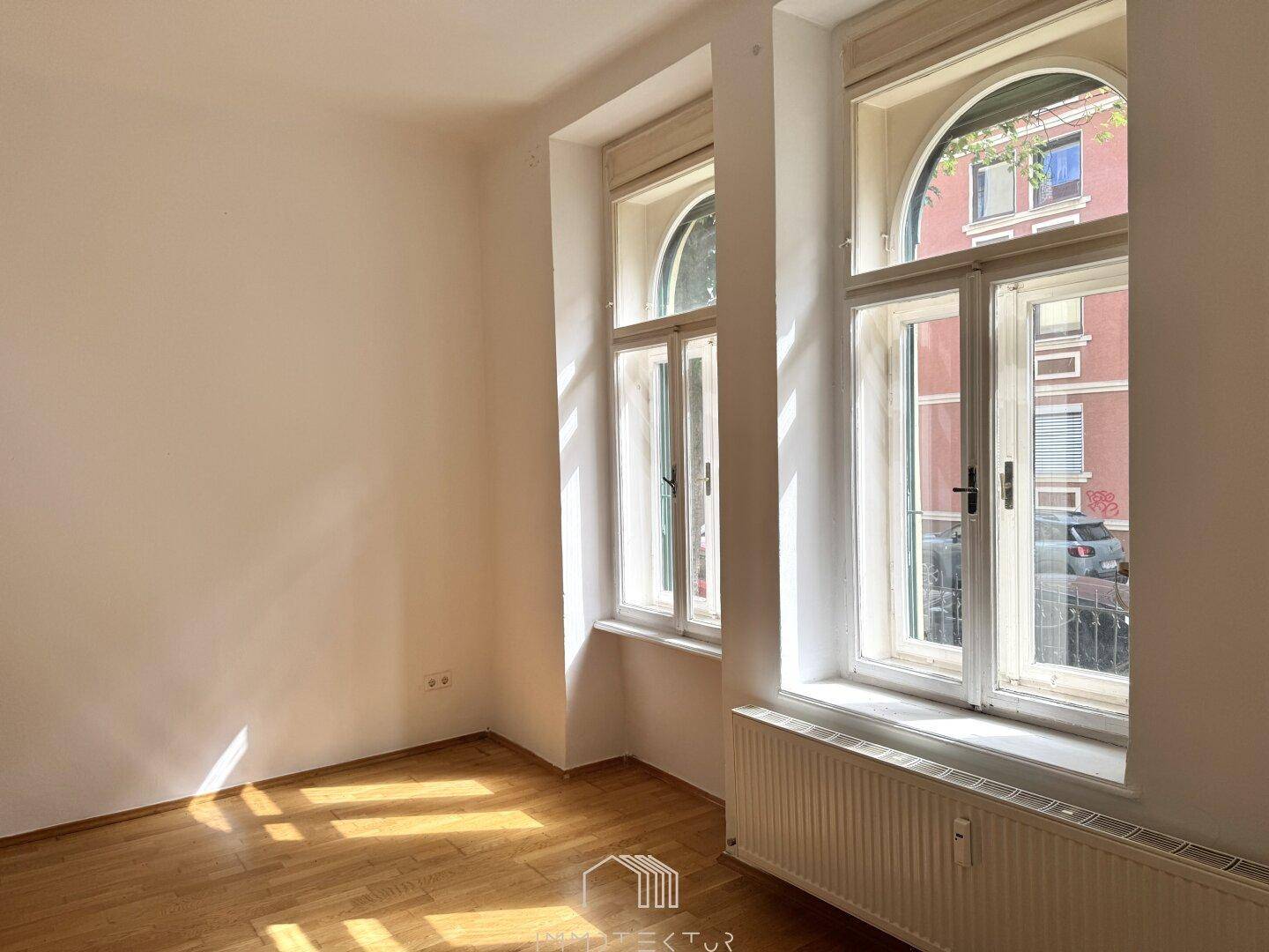 Lichtdurchfluteter Raum mit Parkettboden, weißen Wänden und zwei hohen Fenstern, eines davon mit französischem Balkon.