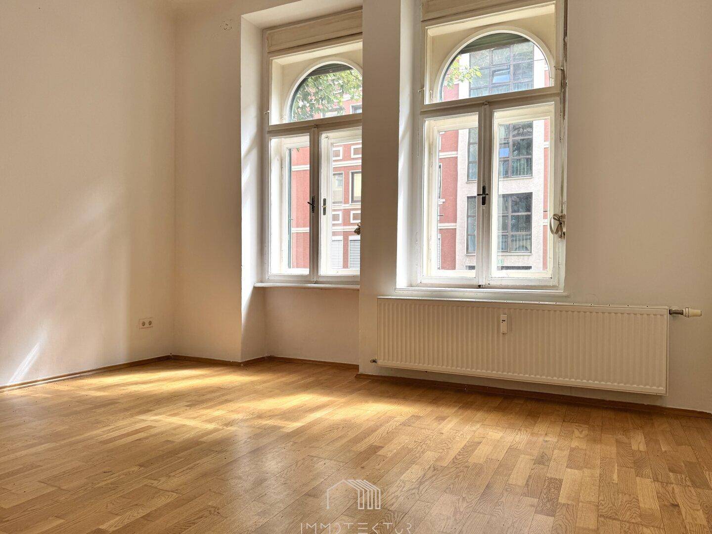 Heller Raum mit Fischgrätparkett, weißen Wänden und zwei großen Bogenfenstern, die viel Tageslicht hereinlassen.