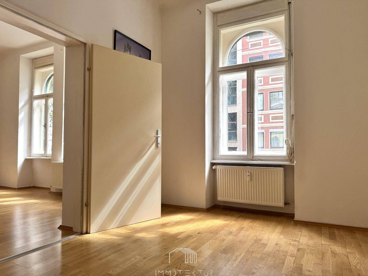 Lichtdurchfluteter Raum mit Holzboden und hohen Fenstern, eine Tür führt in den angrenzenden Nebenraum.