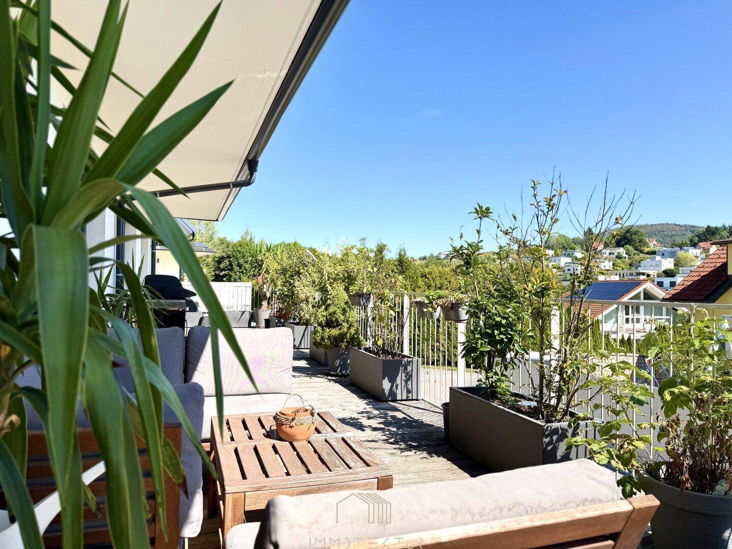 Sonnige Terrasse mit bequemen Gartenmöbeln, vielen Pflanzen und weitem Blick über die grüne Umgebung.