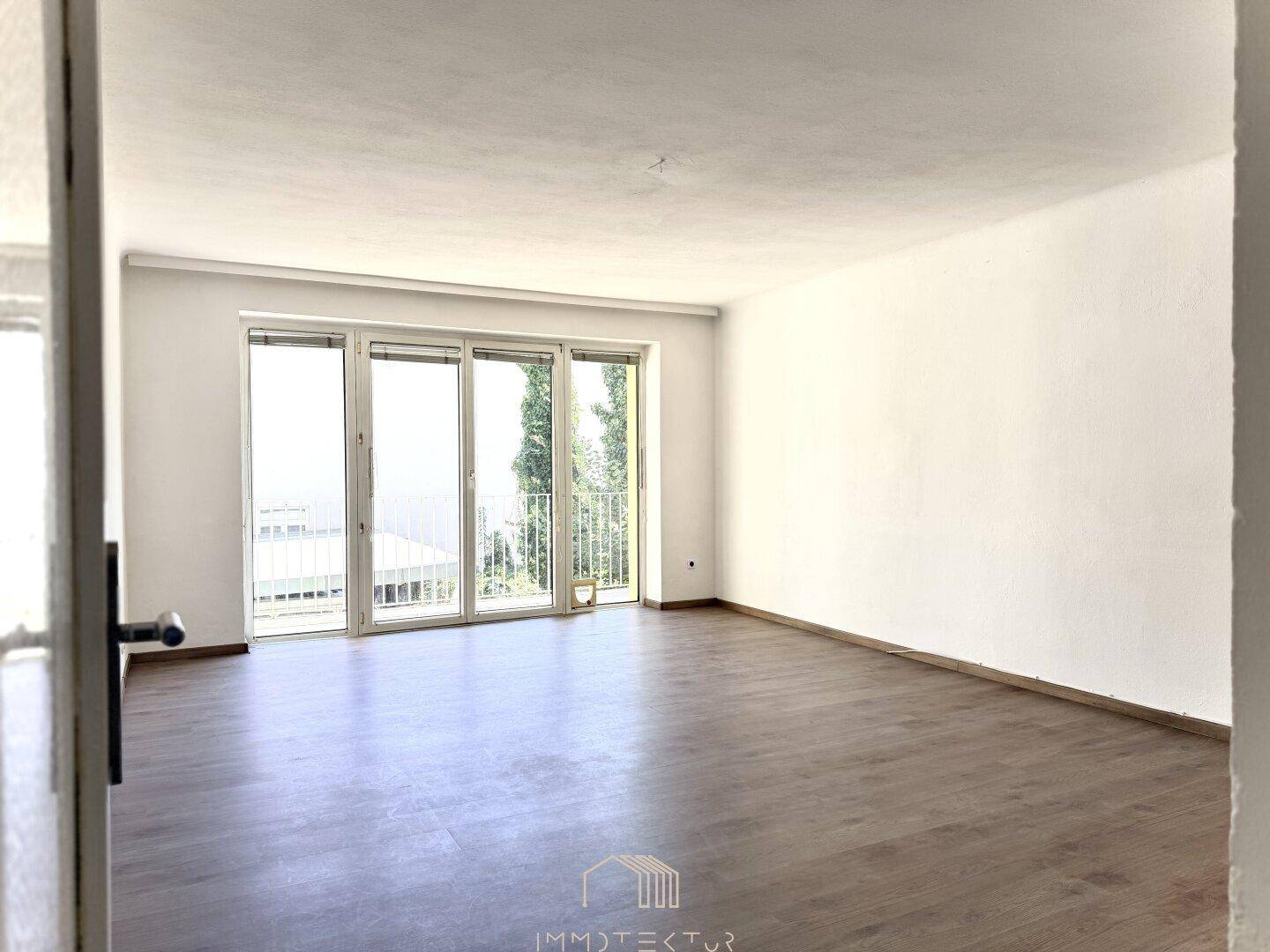 Helles, leeres Wohnzimmer mit Laminatboden und großen Fenstern zum Balkon.