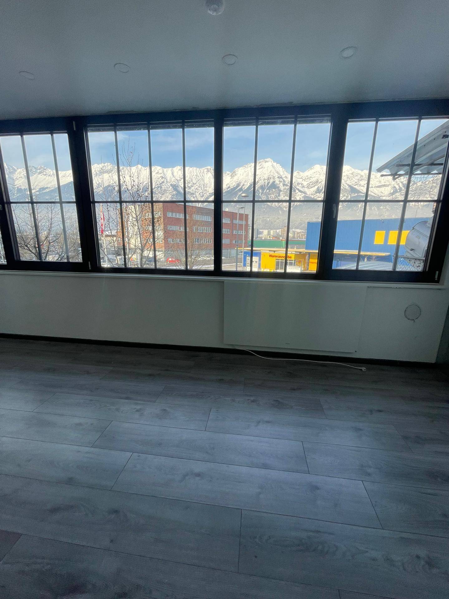 Panoramablick auf schneebedeckte Berge durch große Fenster in einem hellen Innenraum mit Heizkörpern.
