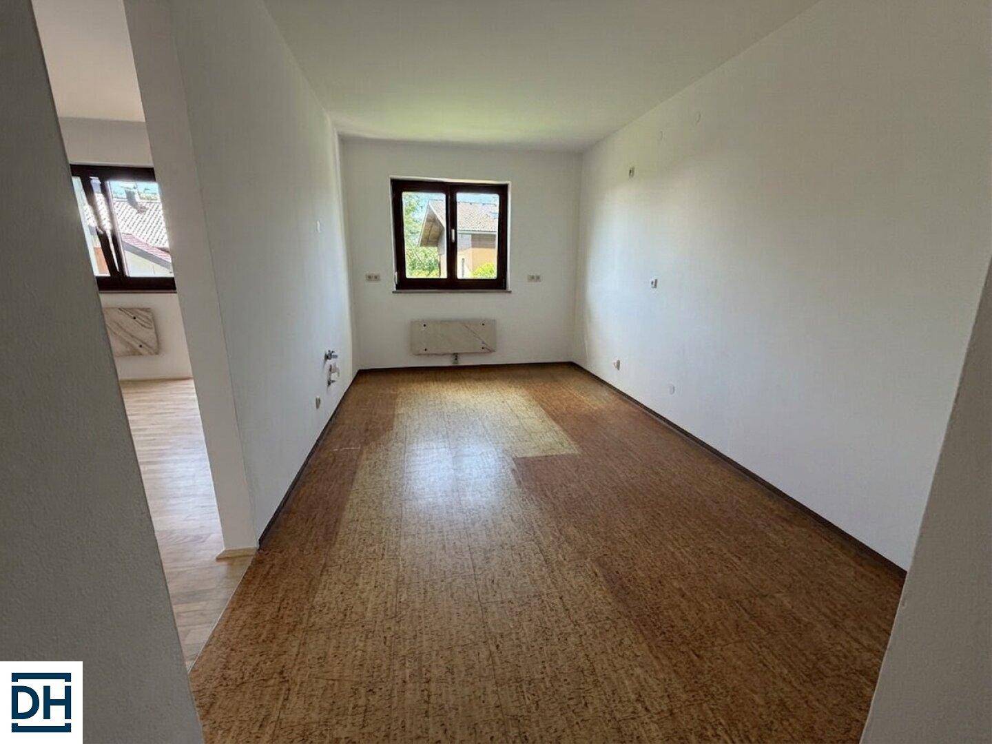 Leerer Raum mit Korkboden, weißen Wänden und einem Fenster, das Tageslicht hereinlässt.