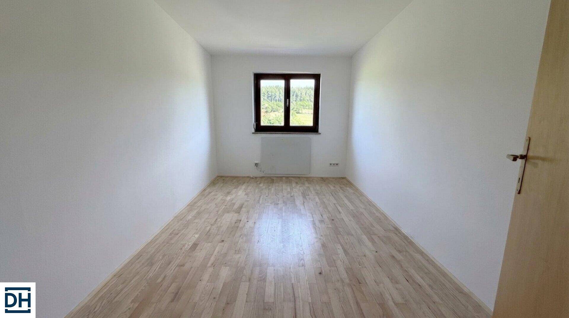 Heller, leerer Raum mit hellem Holzplankenboden, weißen Wänden und einem Fenster mit Blick ins Grüne.