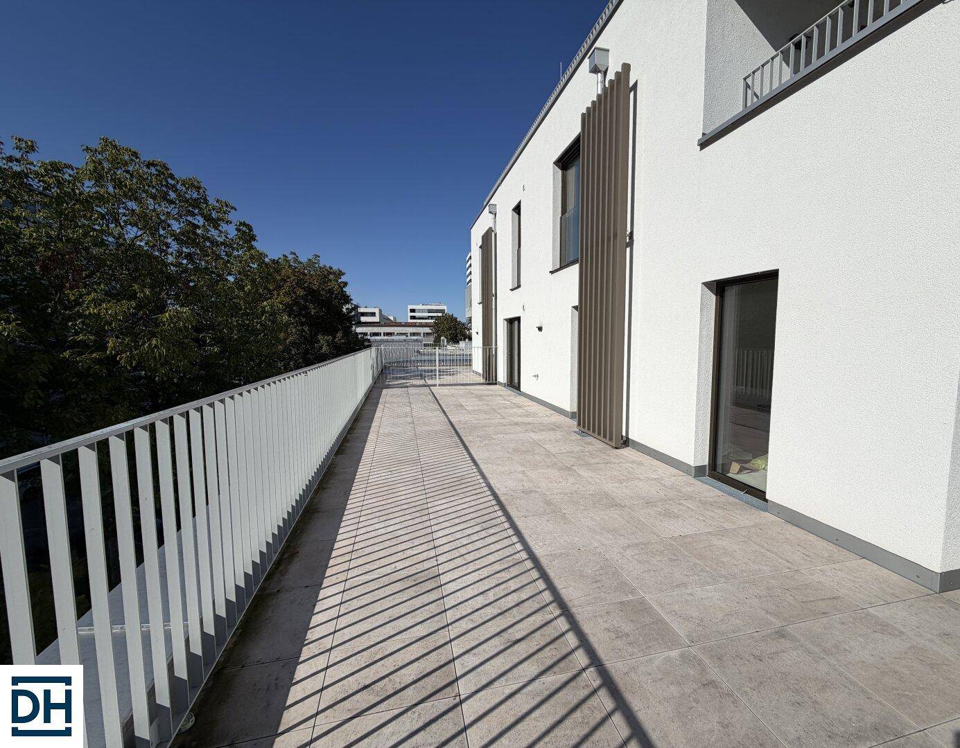 Lange, sonnige Terrasse mit hellem Fliesenboden und weißem Geländer entlang der modernen Fassade.