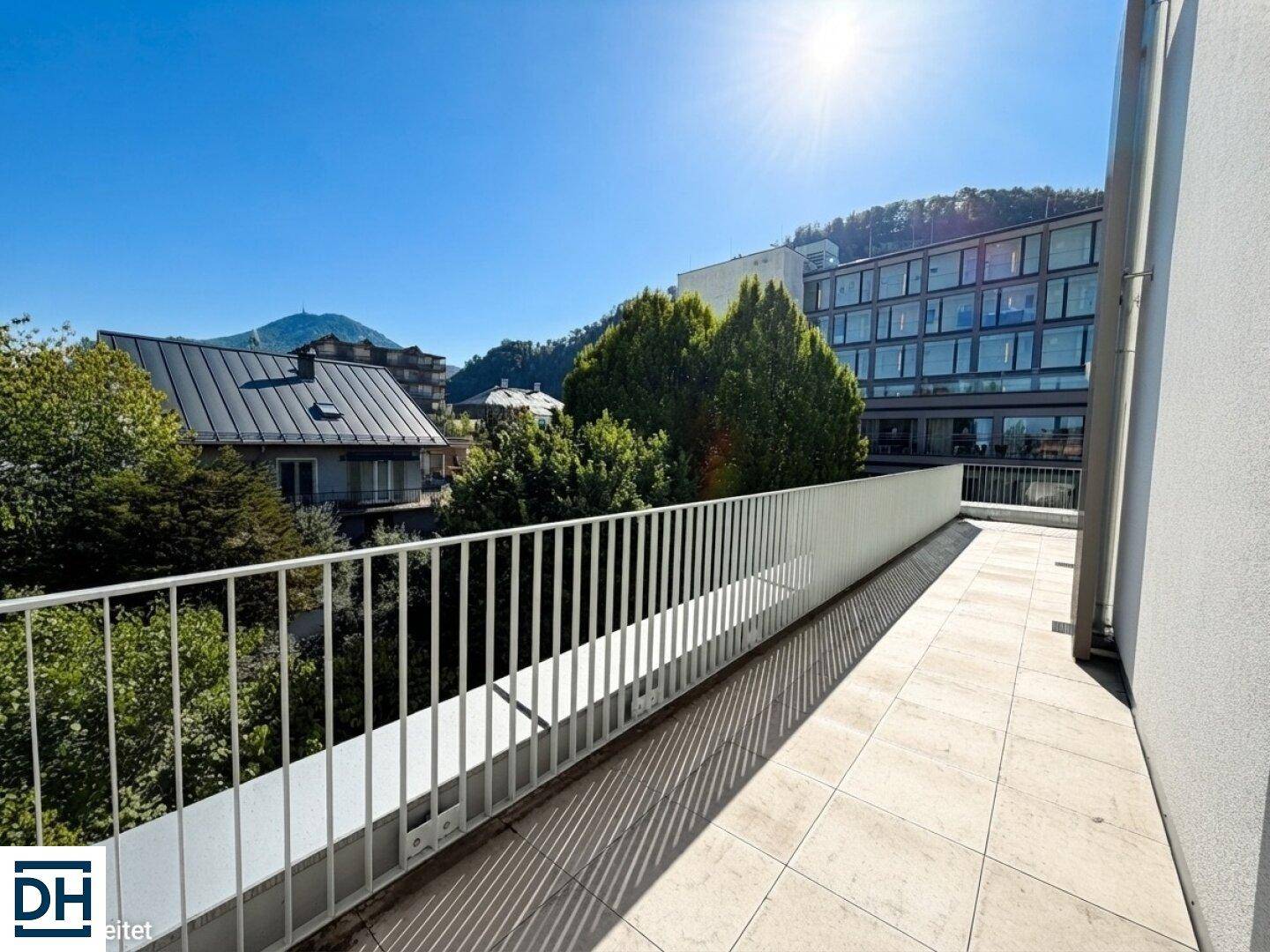 Sonnige Terrasse mit weißem Geländer und Ausblick auf grüne Bäume und die umliegende Bebauung.
