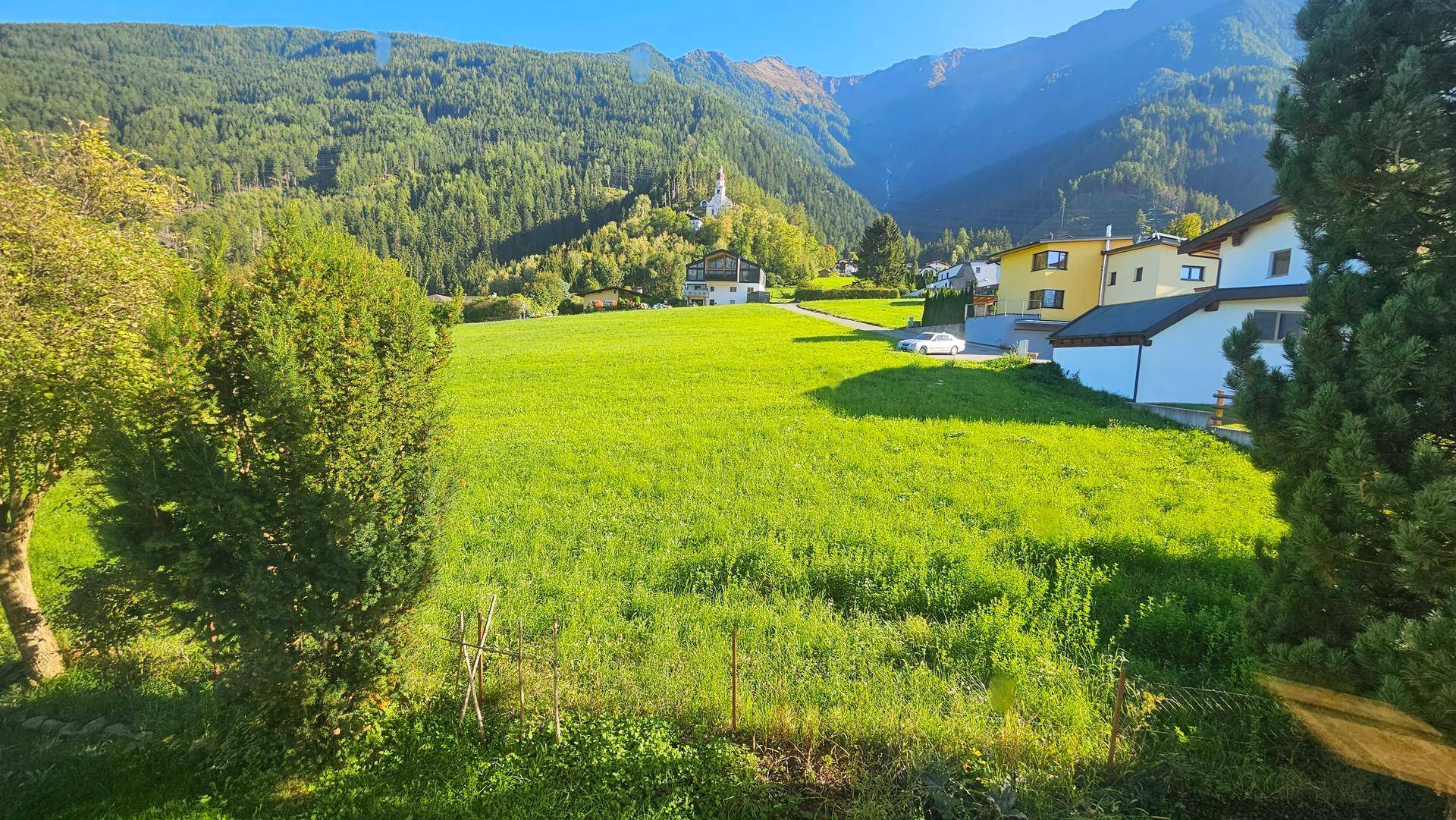 Idyllische Landschaft mit grünen Wiesen, bewaldeten Hügeln und einem Dorf in der Ferne unter blauem Himmel.