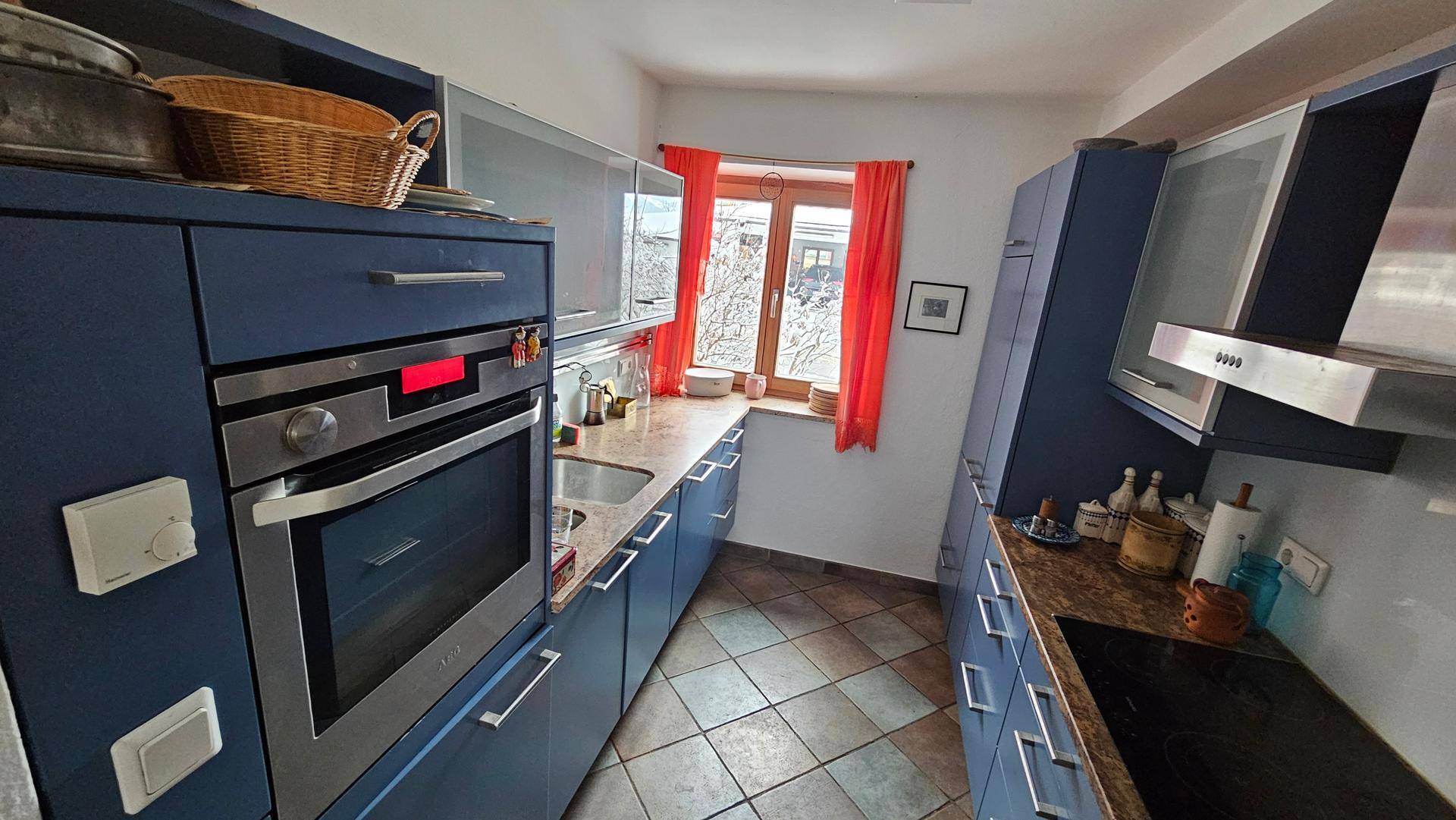 Funktionale Küche mit blauen Schränken, integriertem Backofen und Fenster mit rotem Vorhang.