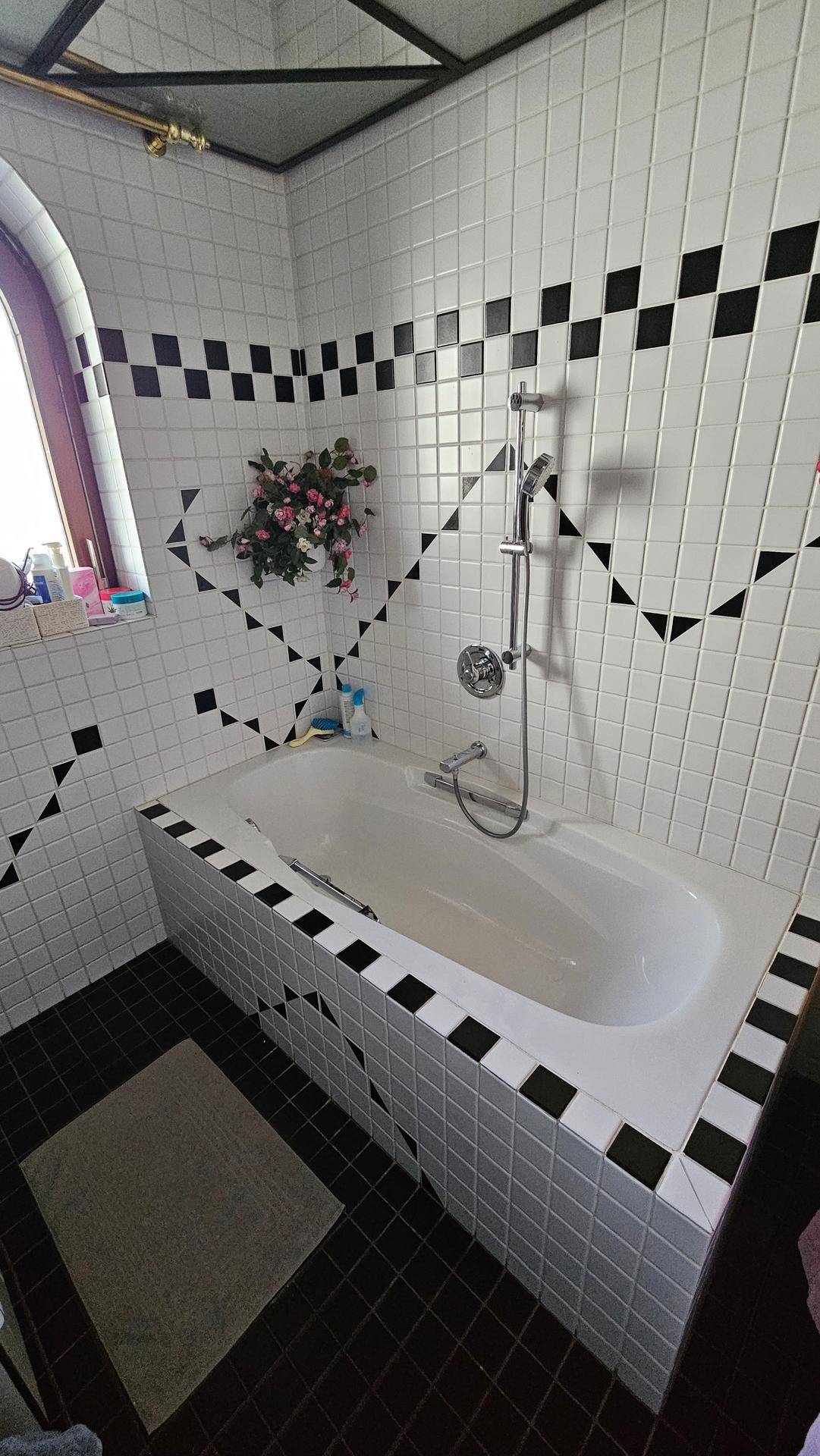 Badezimmer mit Badewanne, Handbrause und charakteristischen schwarz-weißen Fliesen im 80er-Jahre-Stil.