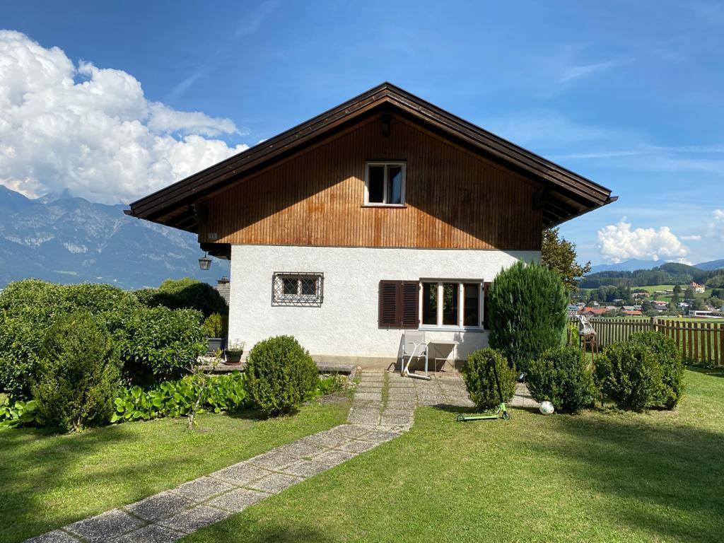 Vorderansicht des Hauses mit gepflegtem Garten, Steinweg und Bergblick unter strahlend blauem Himmel.