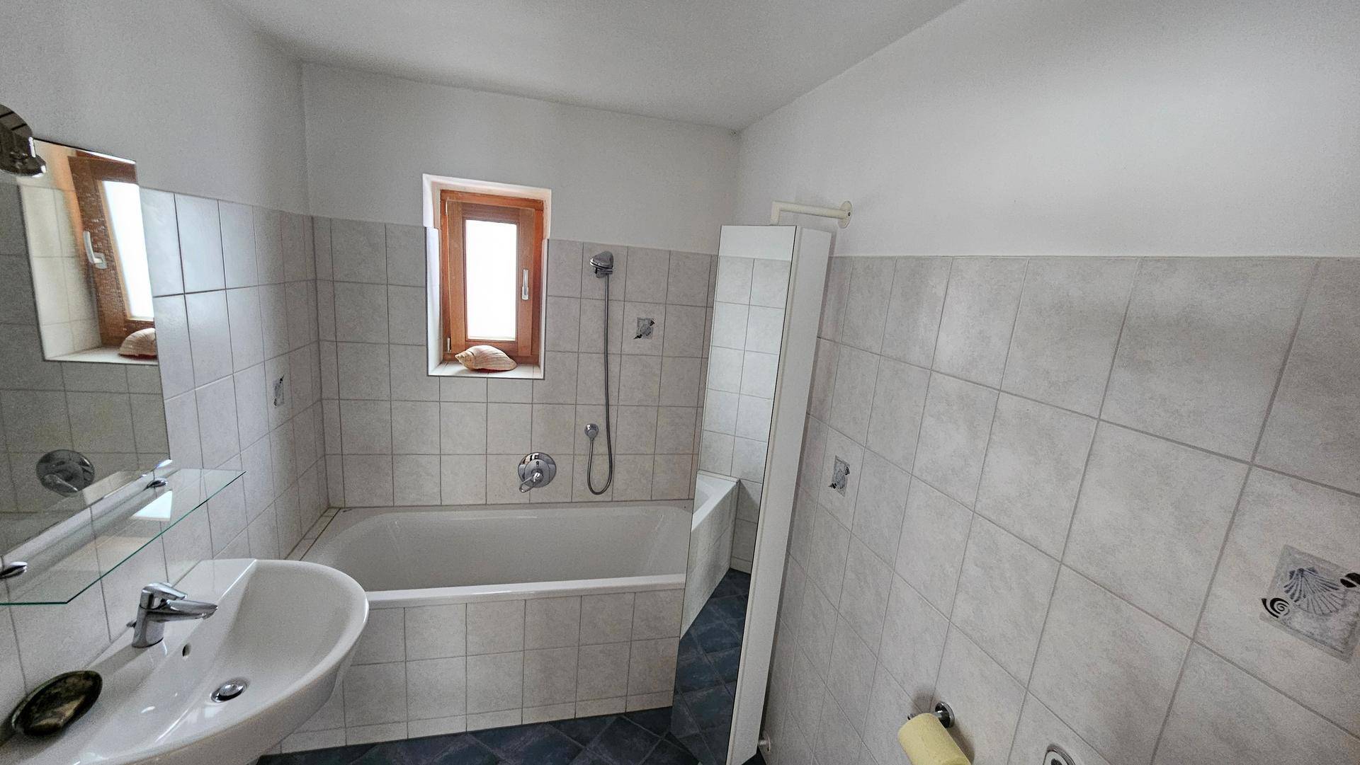 Badezimmer mit Badewanne, Waschbecken und Fenster, hell gefliest.
