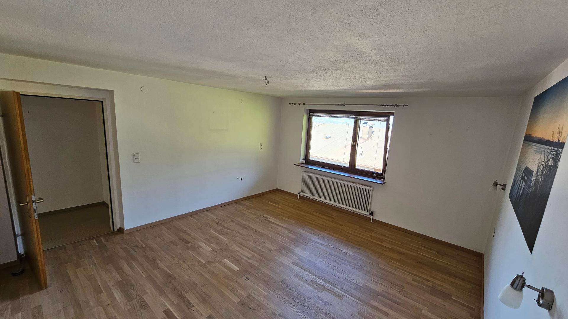 Schlafzimmer mit Holzboden, Heizkörper unter dem Fenster und schlichten weißen Wänden.