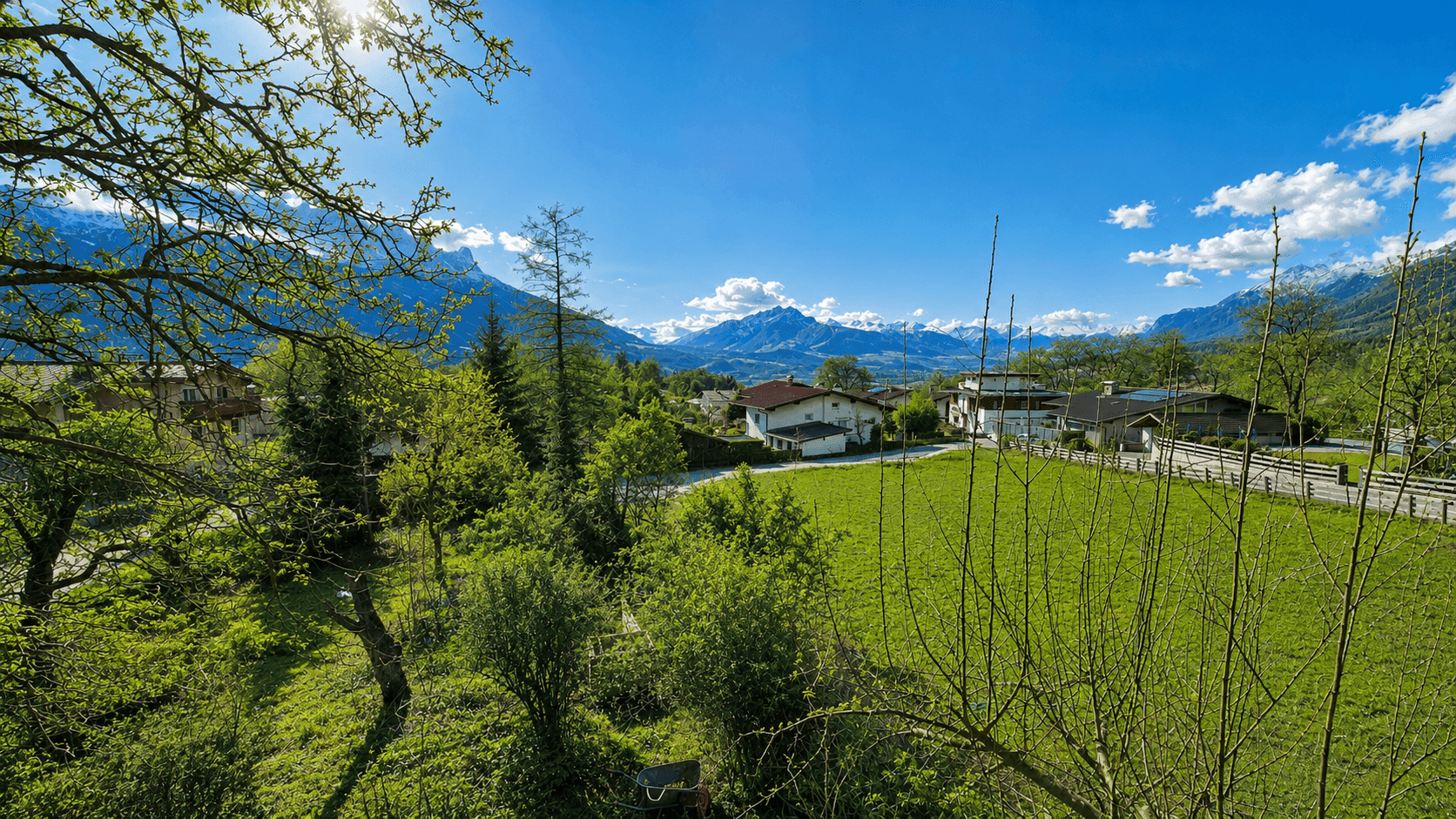 Sonniger Ausblick auf grüne Wiesen, Häuser und die umliegende Berglandschaft mit schneebedeckten Gipfeln.