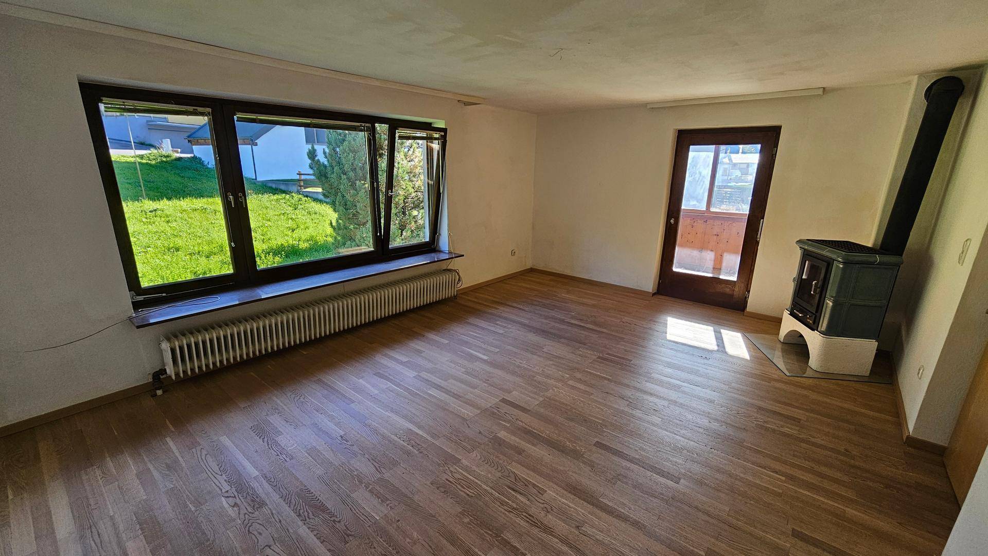 Helles Wohnzimmer mit Holzboden, großen Fenstern und einem traditionellen Kachelofen.