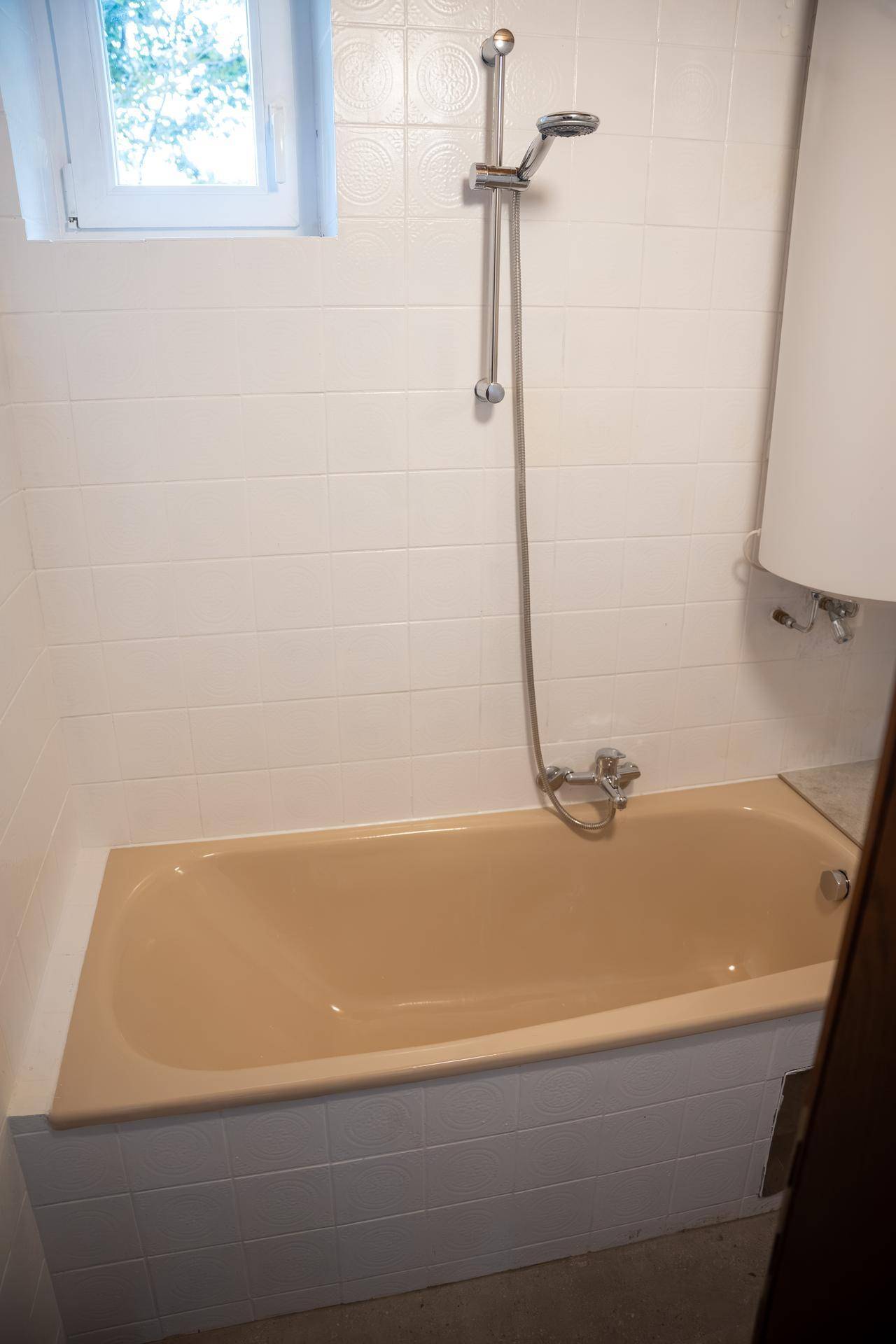 Badezimmer mit beigefarbener Badewanne, Duschvorrichtung und kleinem Fenster.