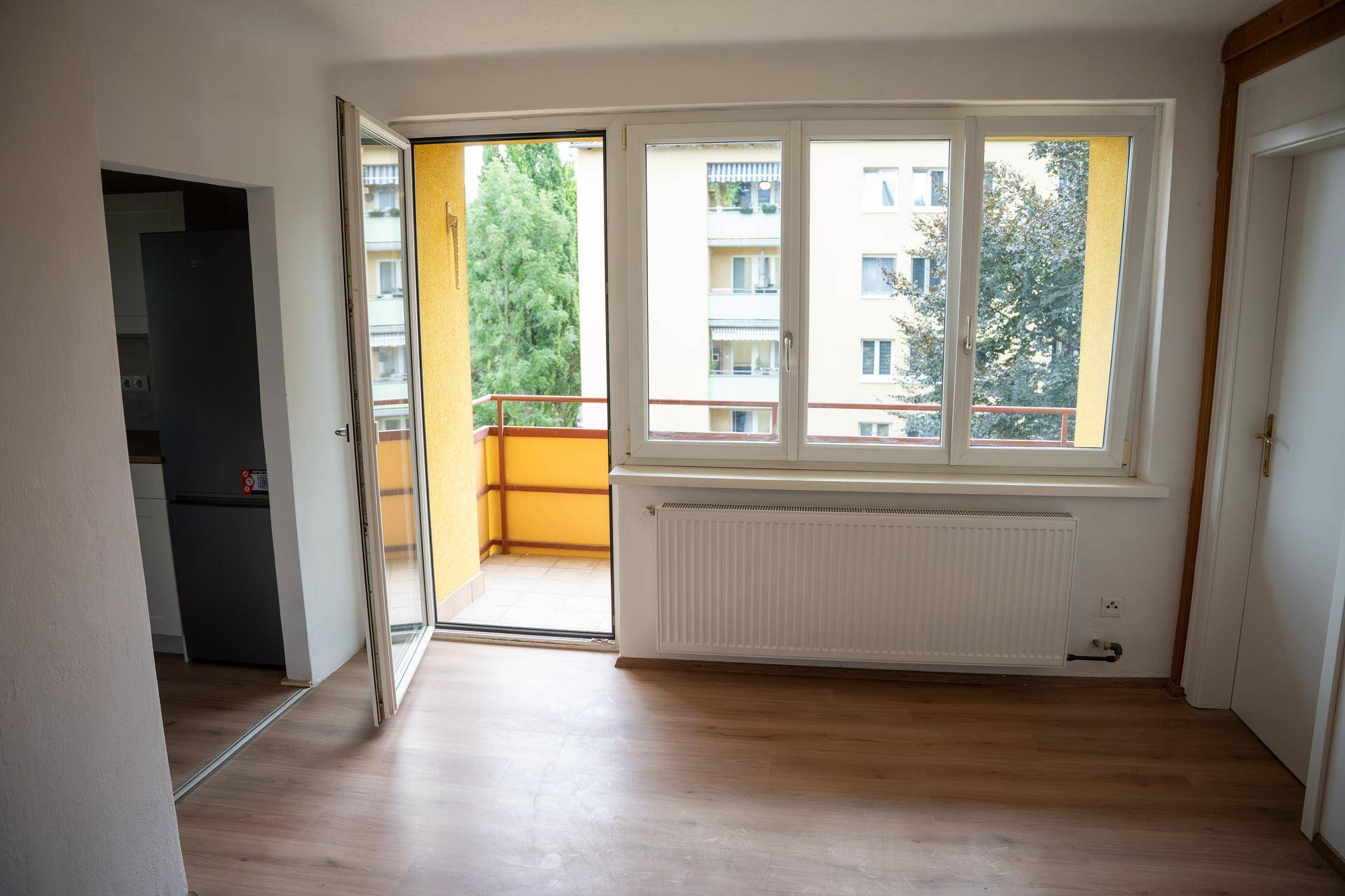 Geräumiges Zimmer mit Laminatboden, Heizkörper und Zugang zum Balkon, mit Blick auf umliegende Gebäude.