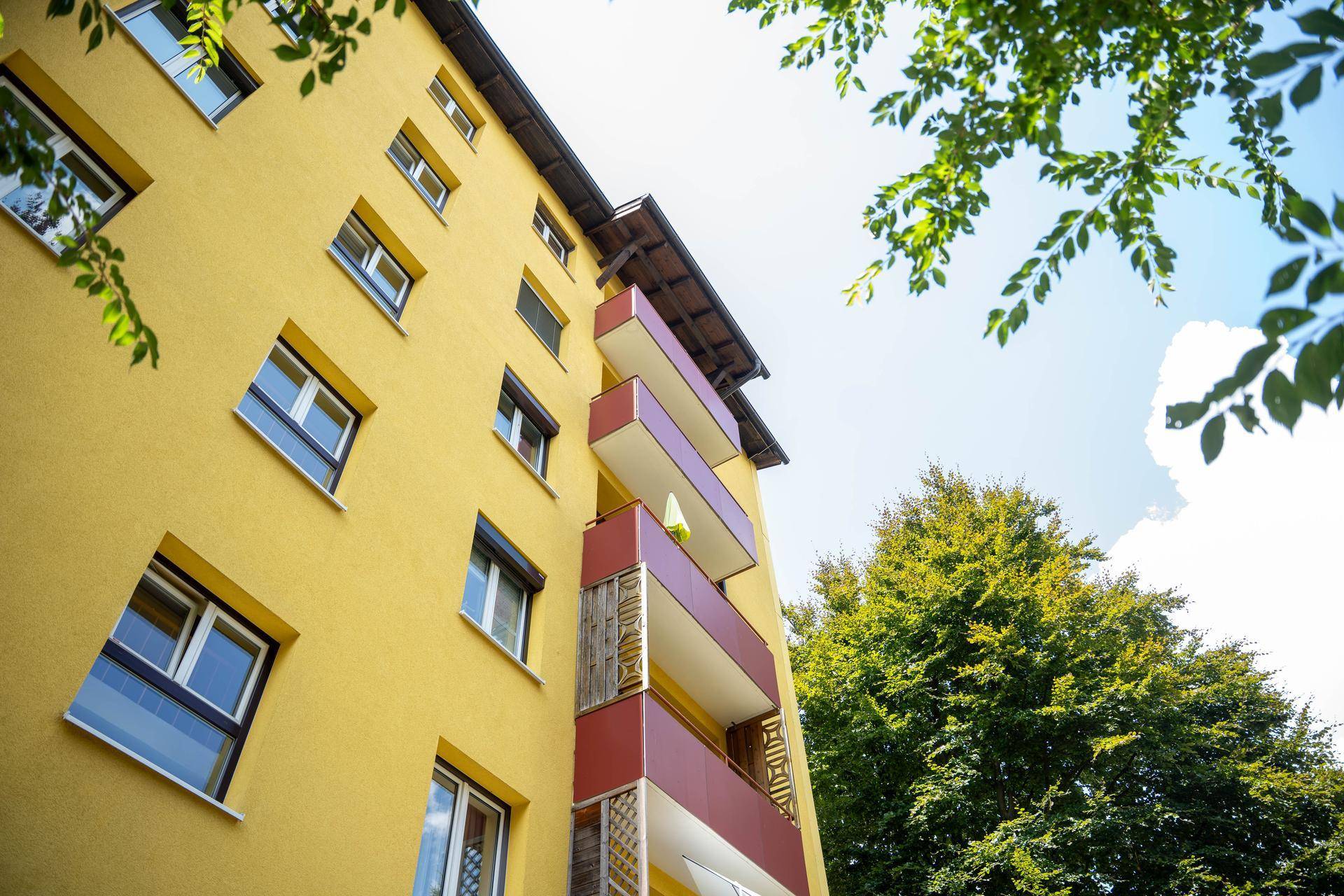 Gelbe Gebäudefassade mit roten Balkonen und grünen Bäumen unter blauem Himmel.