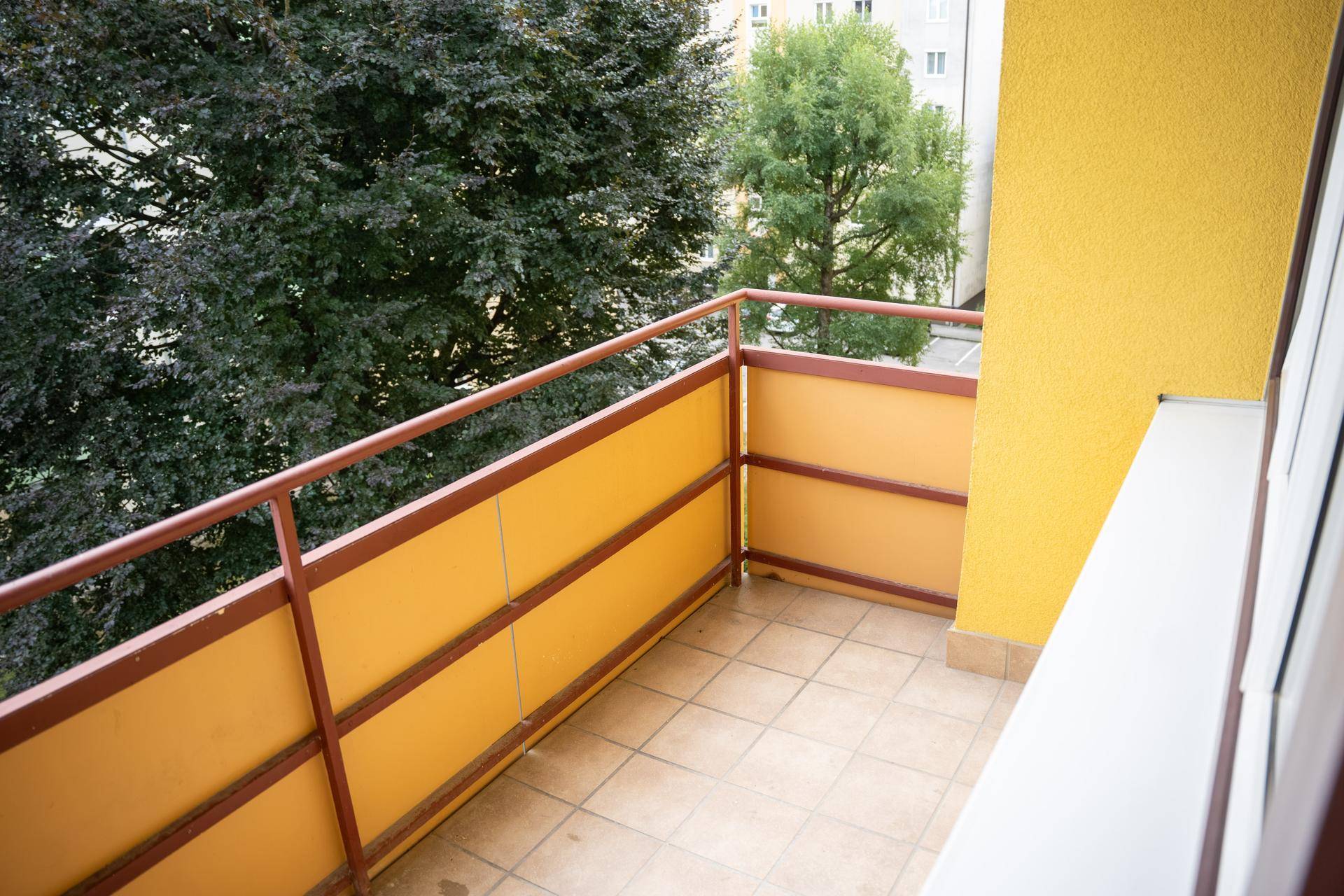 Balkon mit gelbem Anstrich und braunem Geländer, umgeben von grünen Bäumen.