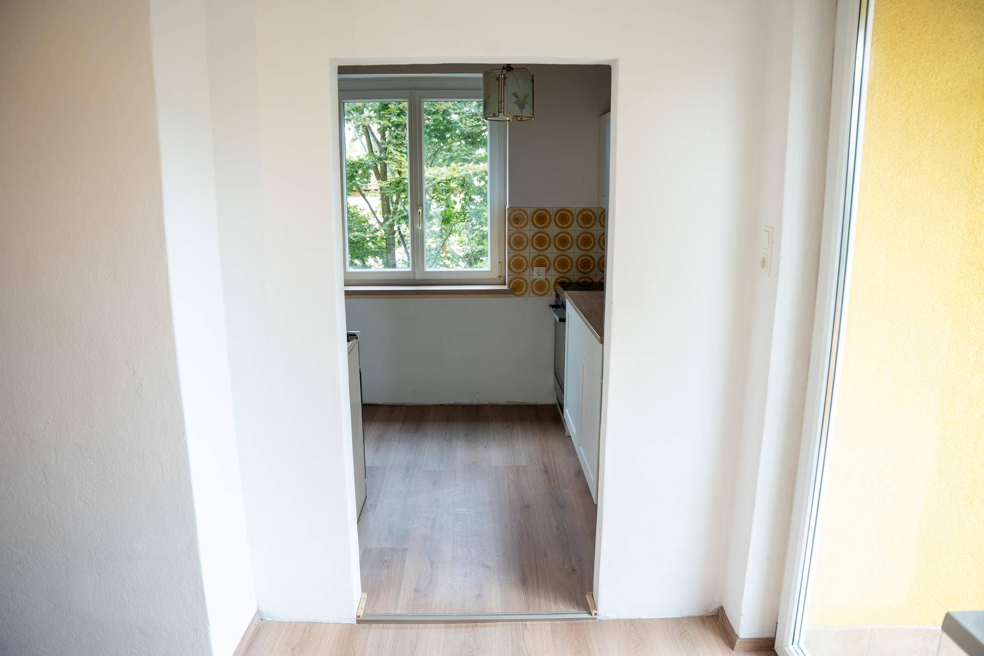 Blick vom Wohnbereich durch einen Durchgang in die kleine Küche mit Fenster und Retro-Fliesen.