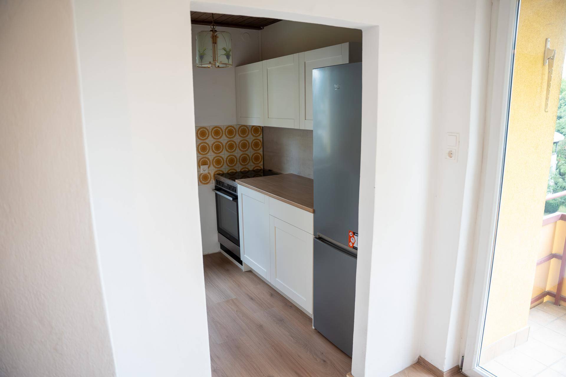 Kleine Küche mit weißen Schränken, Holz-Arbeitsplatte und einem Kühlschrank, sichtbar durch einen Durchgang.