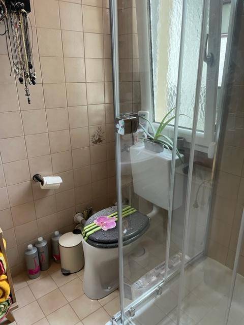 Badezimmer mit beigen Fliesen, Toilette, Duschkabine und einem Fenster.