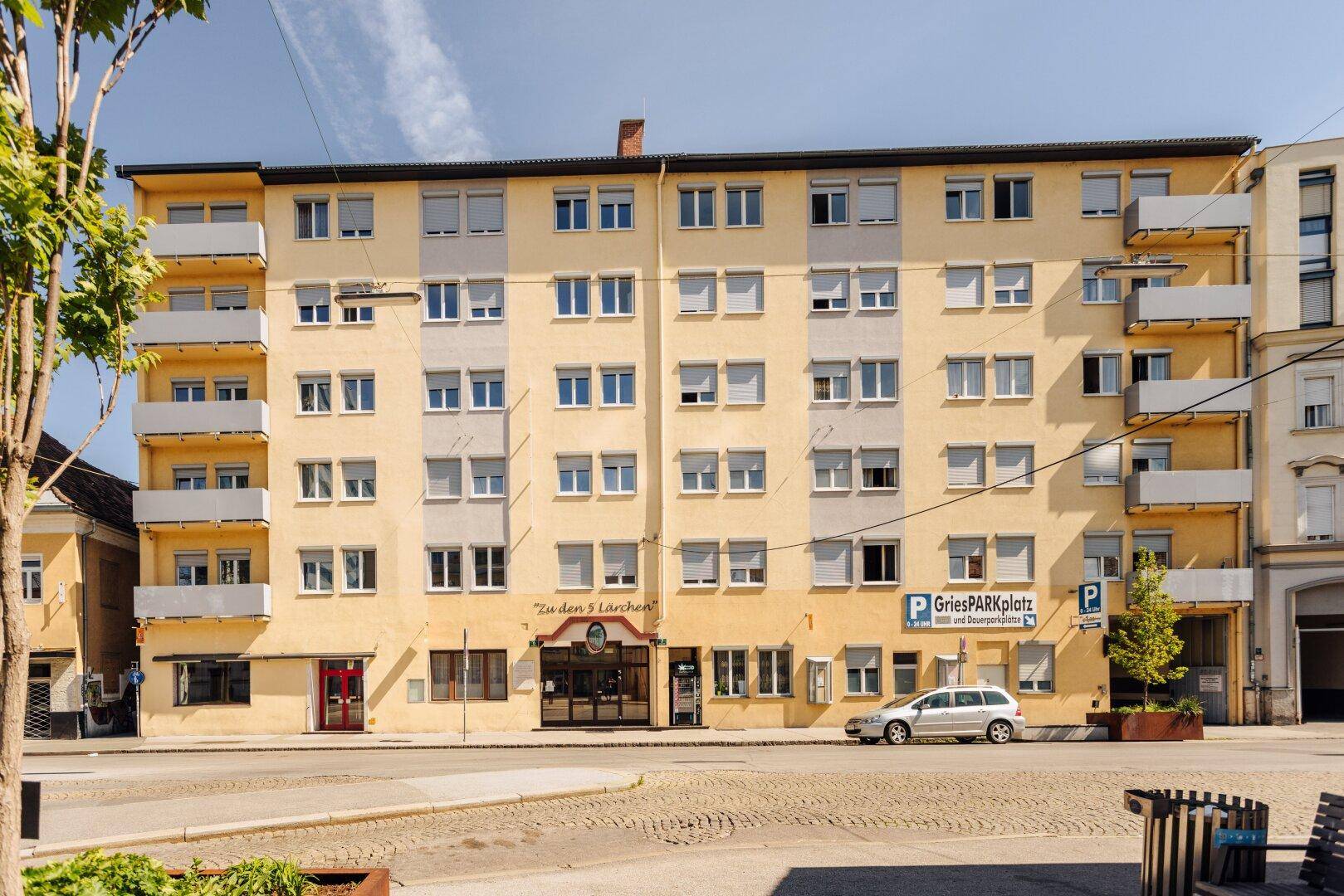 Gesamtansicht des gelben Mehrfamilienhauses mit Balkonen, Fenstern und einem Parkplatzschild.