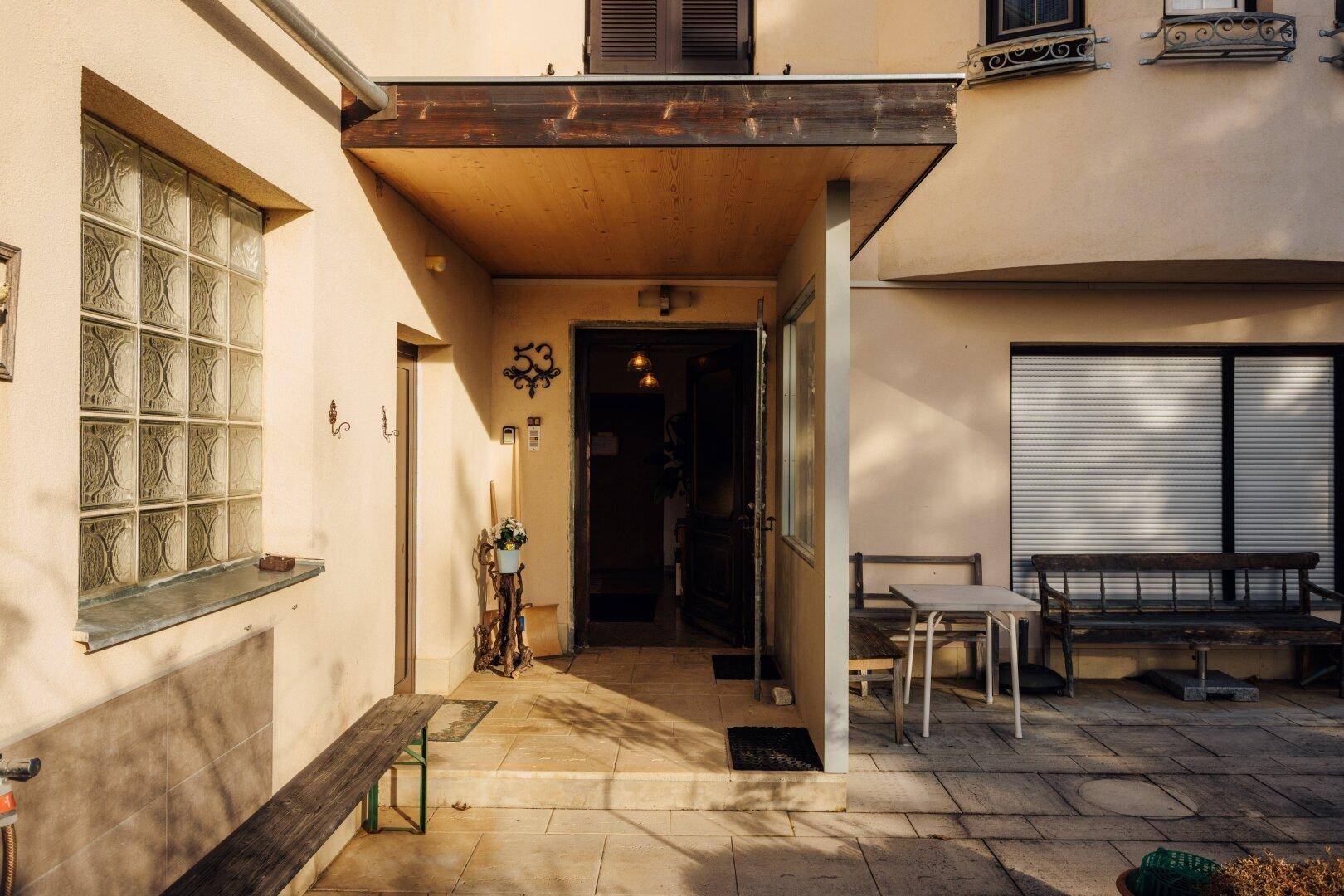 Überdachter Eingangsbereich mit gepflasterter Terrasse und Sitzgelegenheiten im Freien.