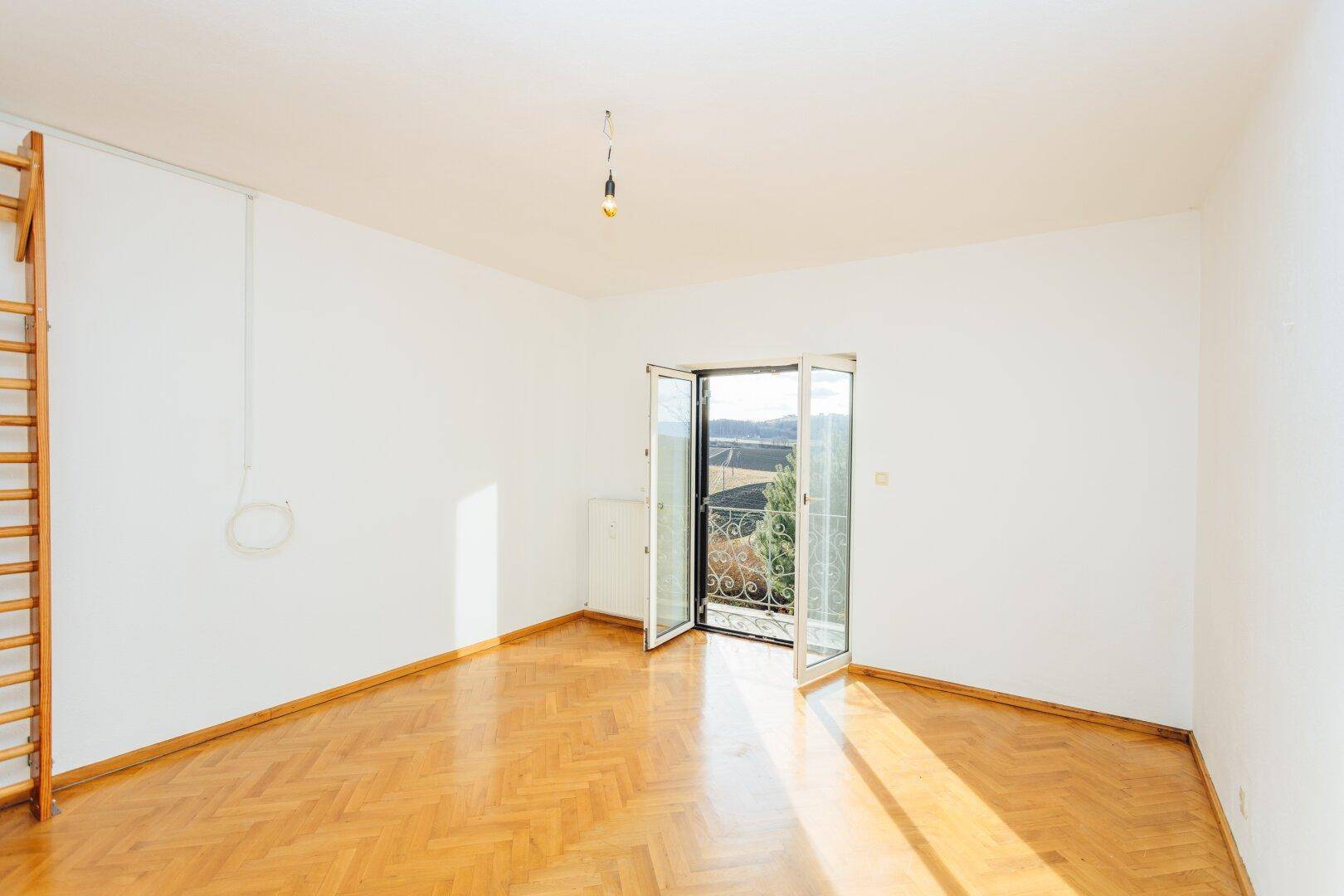 Geräumiges Zimmer mit hellem Fischgrätparkett und Balkontür, die viel Licht hereinlässt.