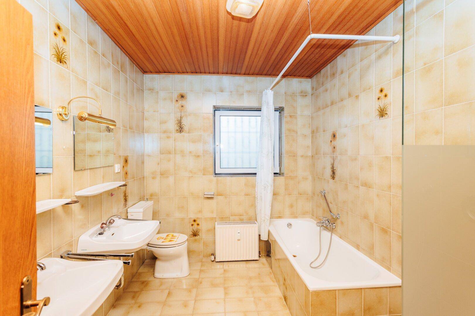 Badezimmer mit beigefarbenen Fliesen, Badewanne, WC, Doppelwaschbecken und Holzdecke.