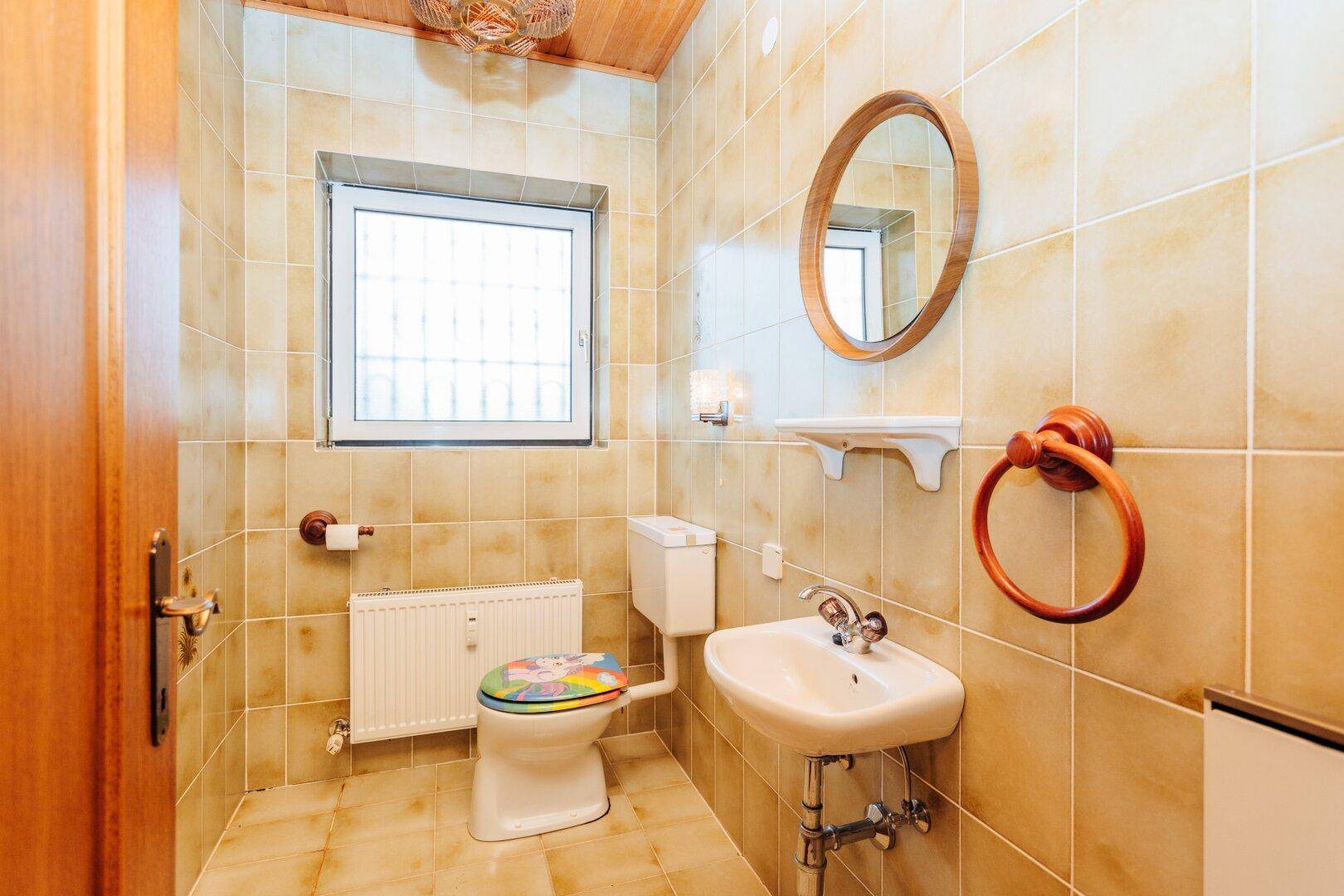 Kleines Badezimmer mit beigefarbenen Fliesen, Fenster, WC und Waschbecken.