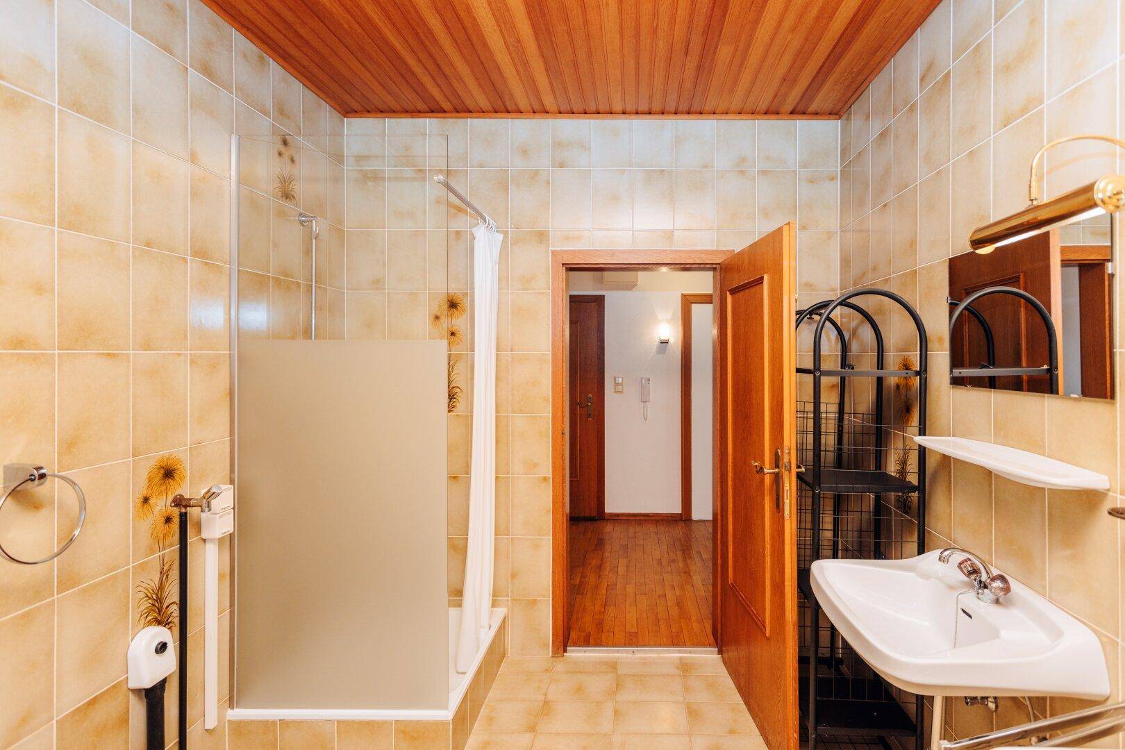 Badezimmer mit beigefarbenen Fliesen, Duschbereich, Waschbecken und Holzdecke.