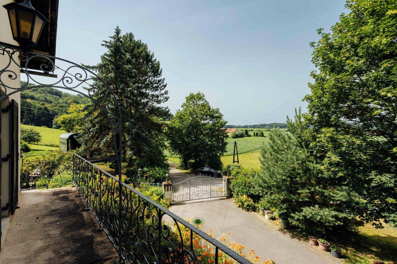 Weitläufiger Blick vom Balkon über die grüne Landschaft und die Zufahrt des Anwesens.
