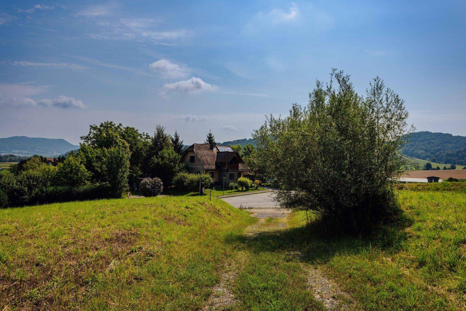 Schotterweg führt zu einem Haus mit markantem Dach, umgeben von Grünflächen und Hügeln.