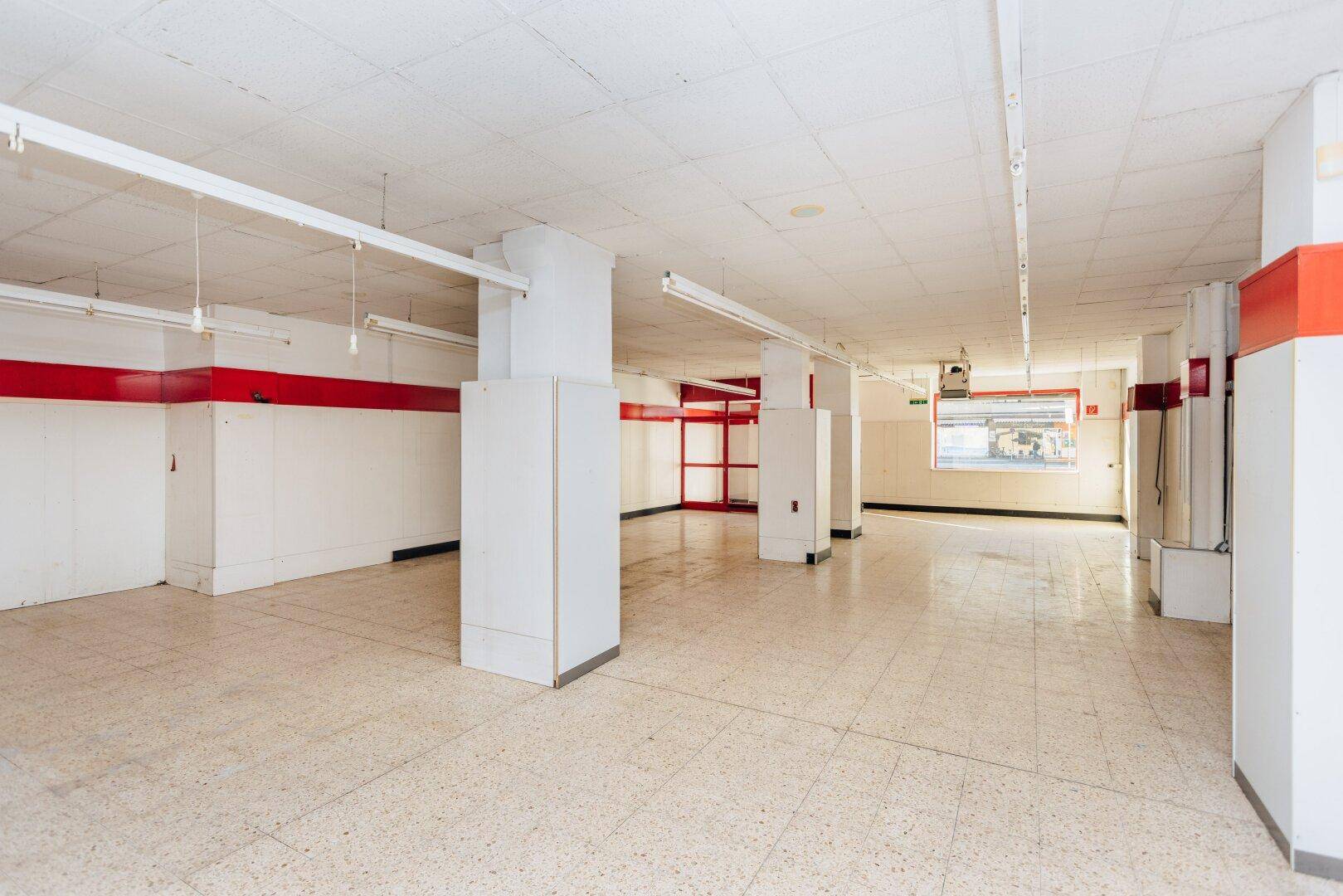 Weitläufiger, leerer Ladenbereich mit Säulen, Fliesenboden und roten Akzenten an den Wänden.