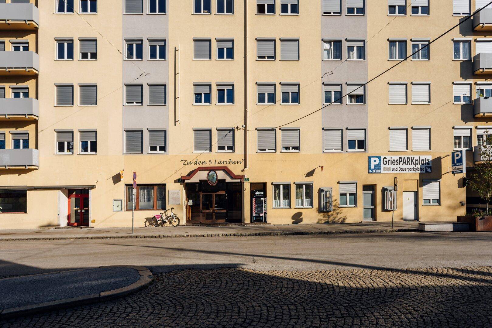 Mehrstöckiges Gebäude mit gelber Fassade, vielen Fenstern und Rollläden an einer Straße.