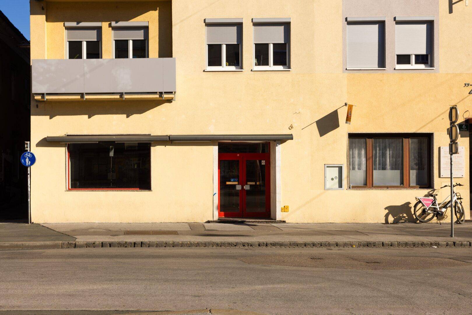 Gelbe Fassade eines Gebäudes mit roten Eingangstüren und Fenstern mit Rollläden.