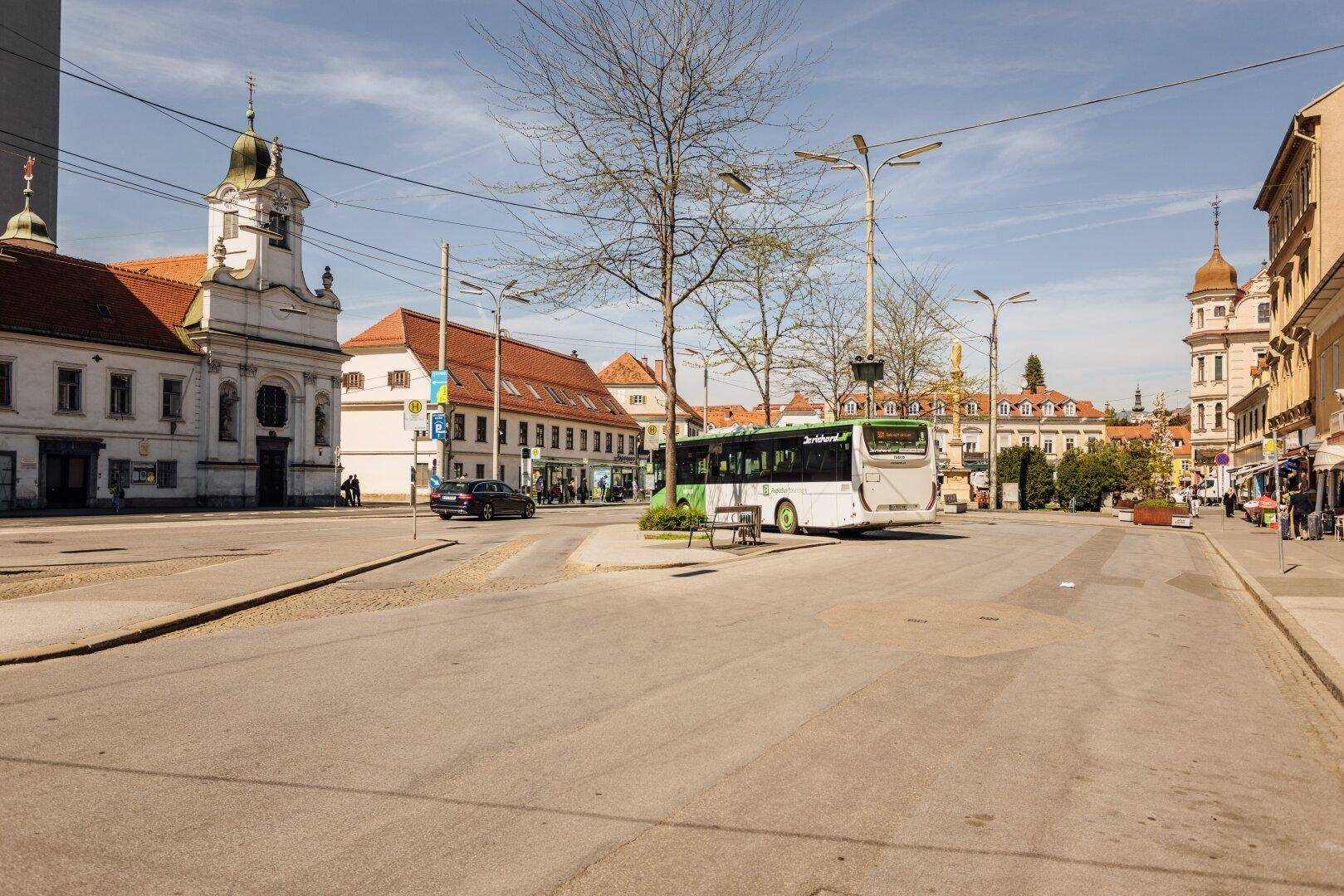 Stadtansicht mit breiter Straße, einem Bus und historischen Gebäuden unter blauem Himmel.