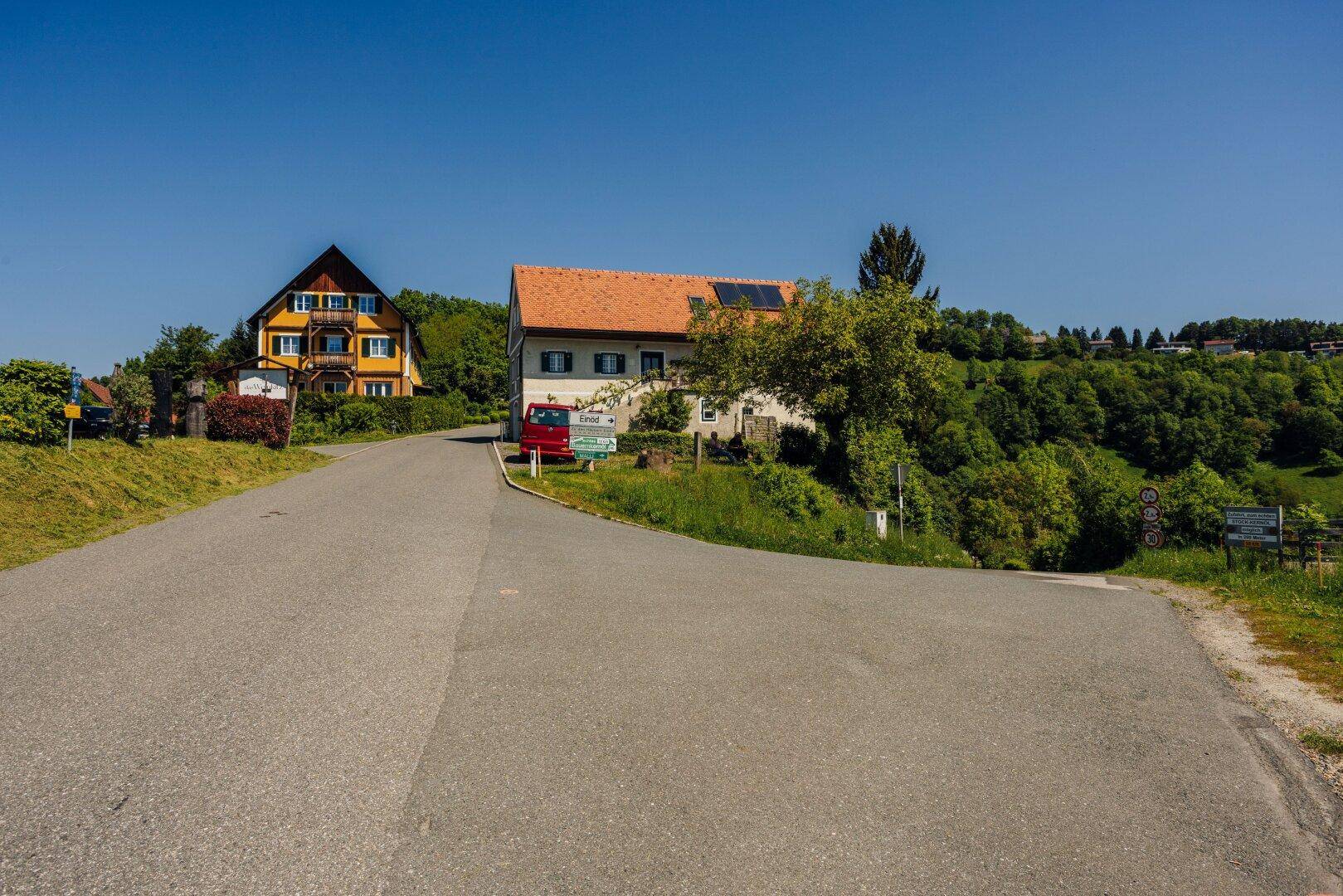 Zwei Häuser an einer ländlichen Straßenkreuzung, umgeben von grünen Hügeln unter blauem Himmel.