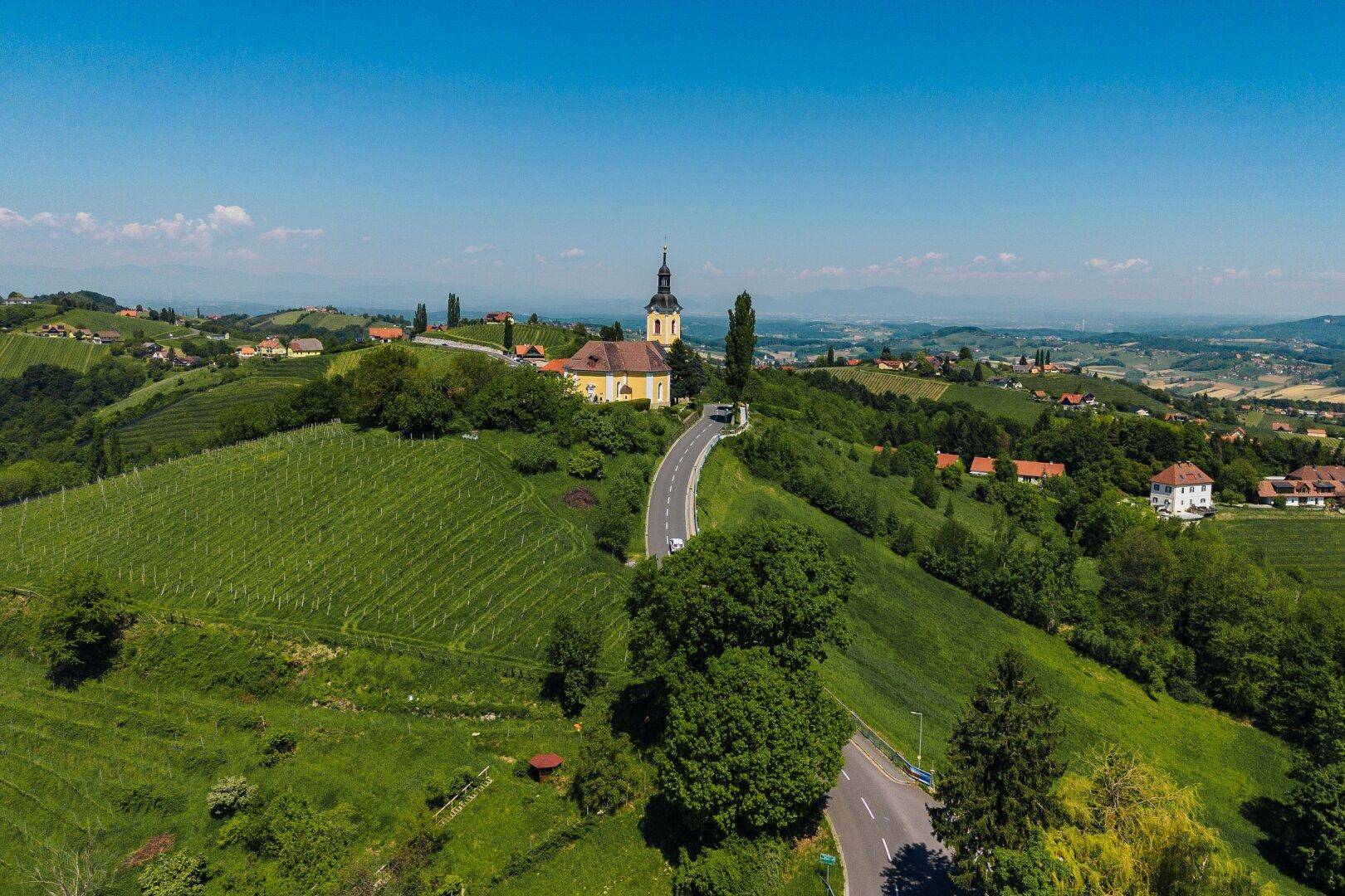 Luftaufnahme einer Kirche auf einem Hügel, umgeben von Weinbergen und einer kurvigen Straße, mit weitem Ausblick.