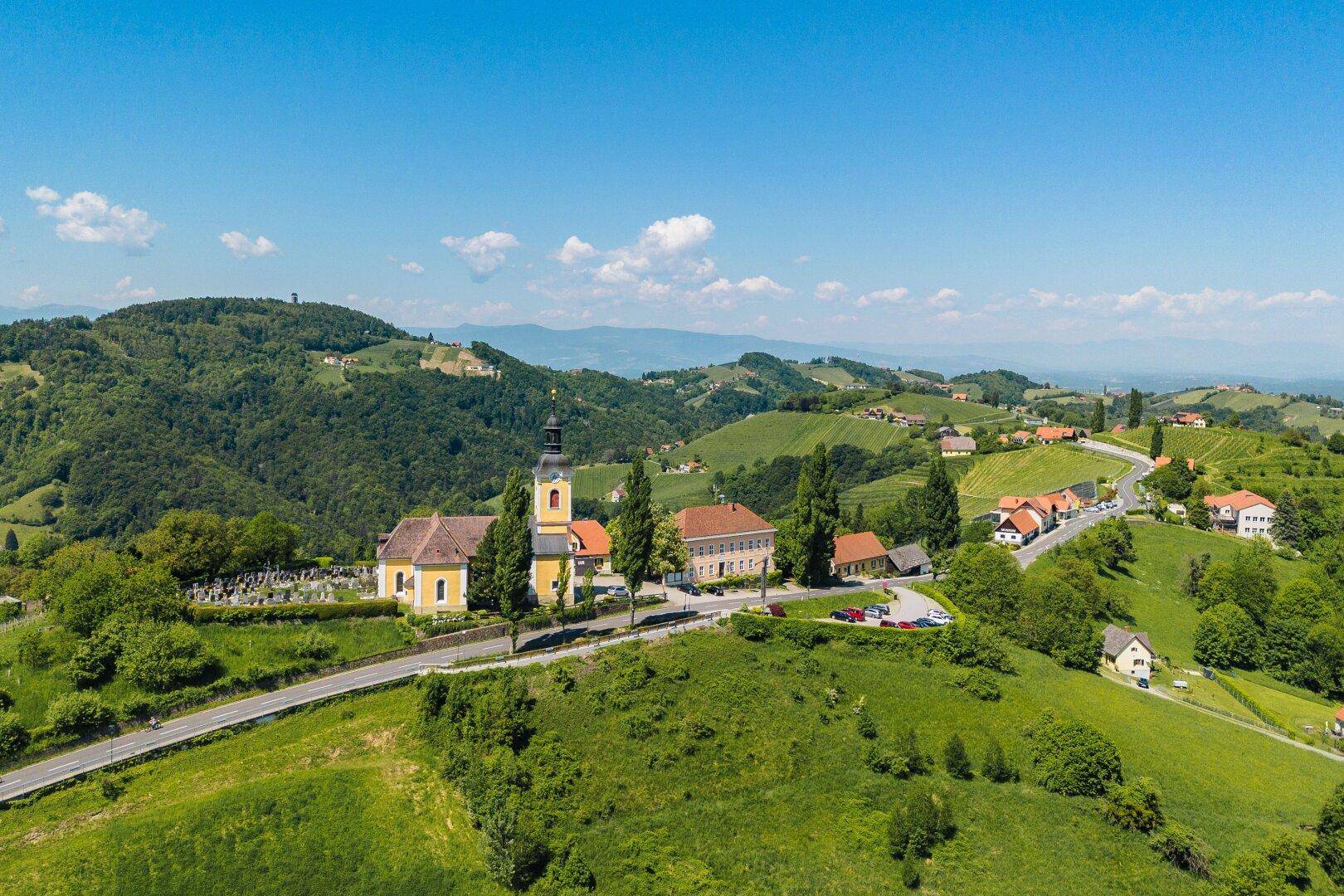 Luftaufnahme eines Dorfes mit Kirche und Gebäuden, eingebettet in grüne Hügel und Weinberge unter blauem Himmel.